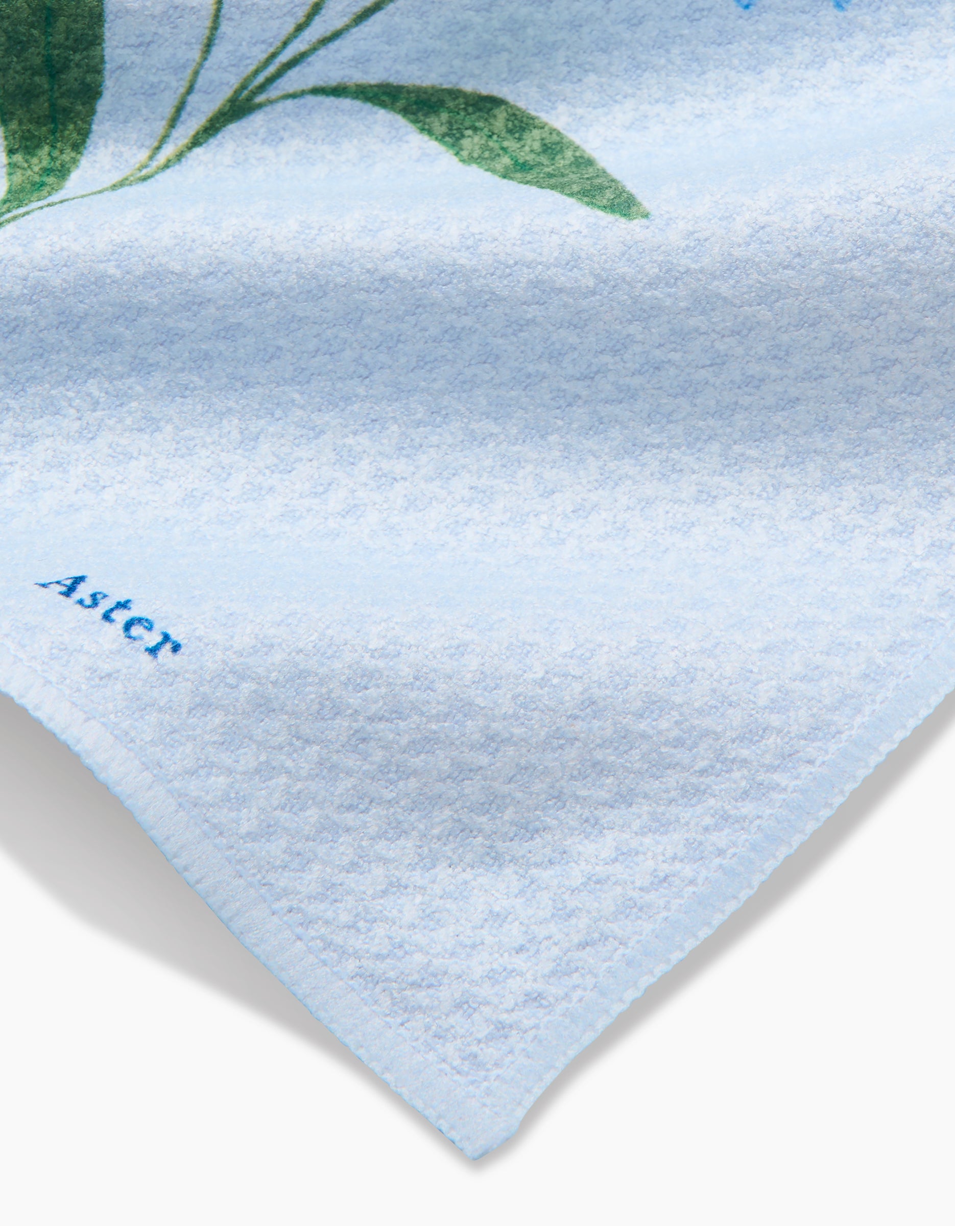 A close-up of Linda Fox’s September Aster dishcloth shows super-absorbent light blue Geoweave fabric with green leaf patterns and “ASTER” printed in blue near the corner.