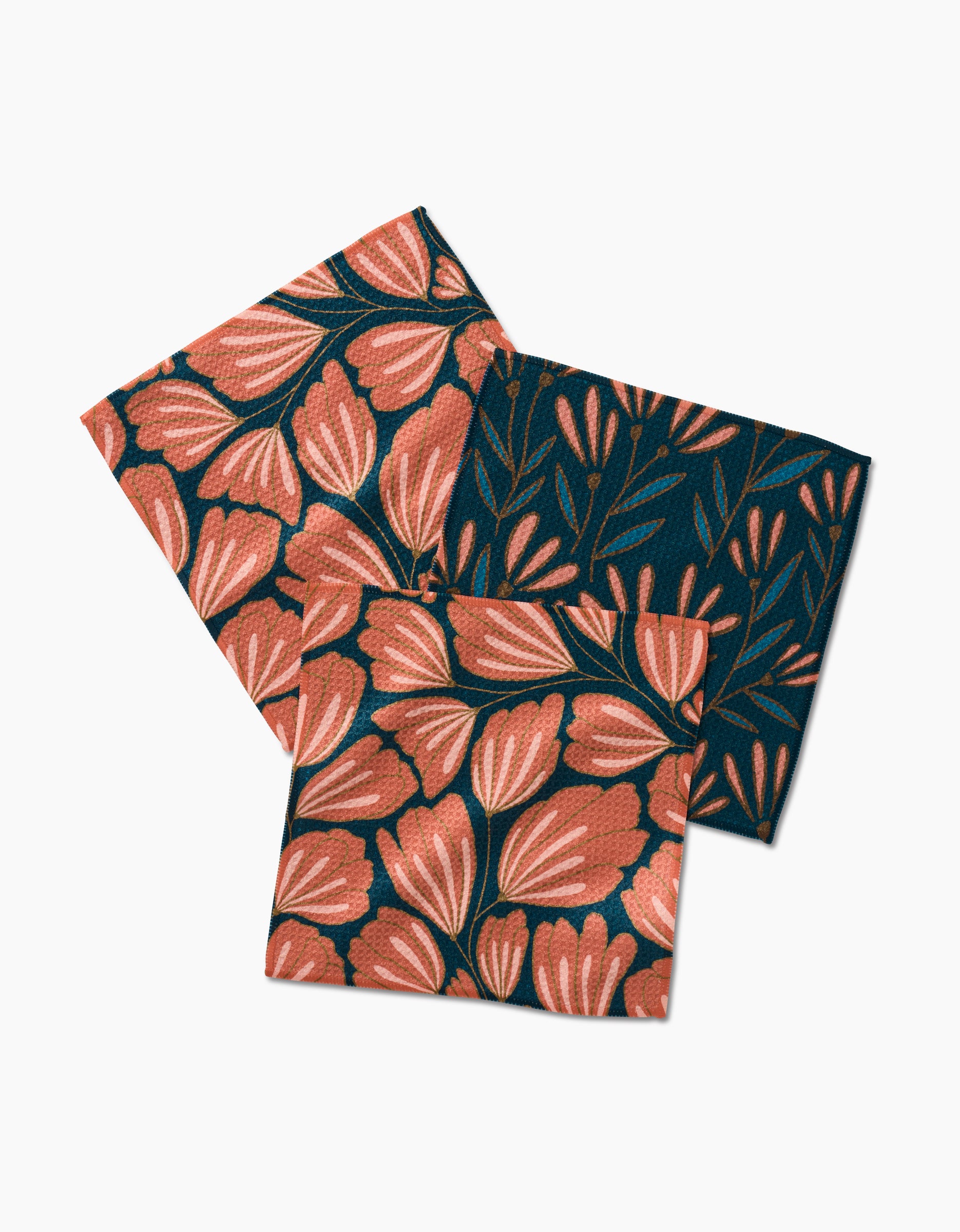 Two Sheila Flower napkins by Amy MacCready with coral-pink floral patterns and one with a dark background and coral-pink leaves, all arranged overlapping on a white surface.