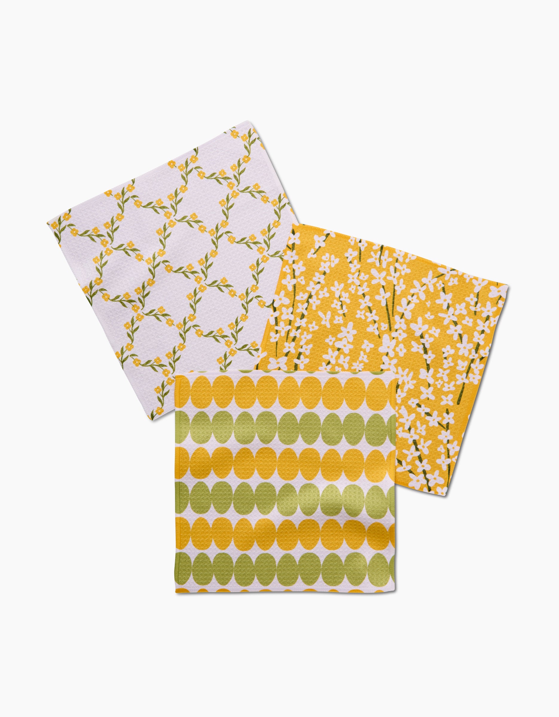 Kayla Anns Signs of Springtime features three Geoweave cloths with lively patterns—leaf vines, white flowers on yellow, and green-yellow circles—overlapping on a white surface.