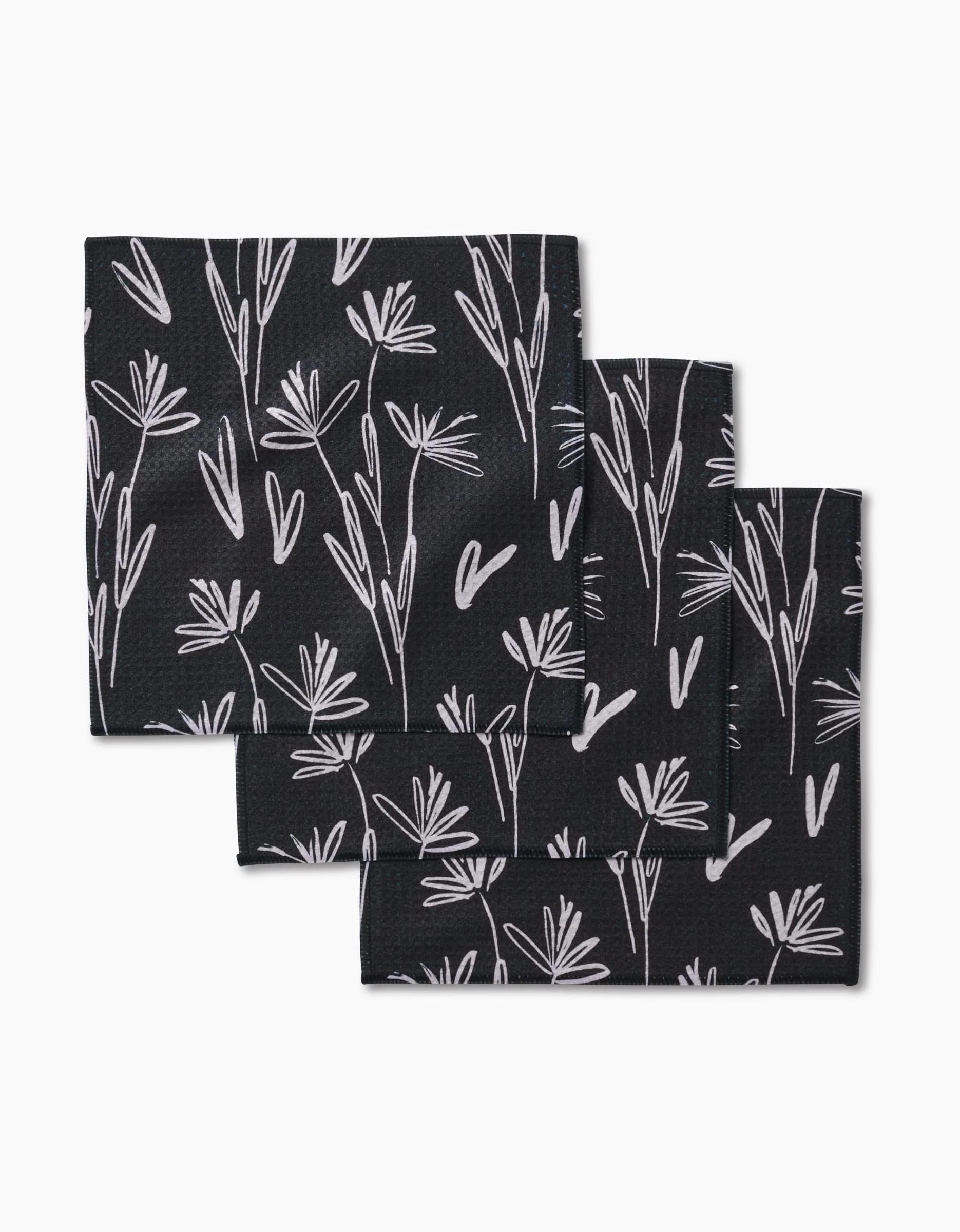 Three Sketchbook Dusk napkins by Toboggan Avenue, crafted from recycled black cloth and featuring a white minimalist floral pattern, are stacked slightly offset on a plain white background.