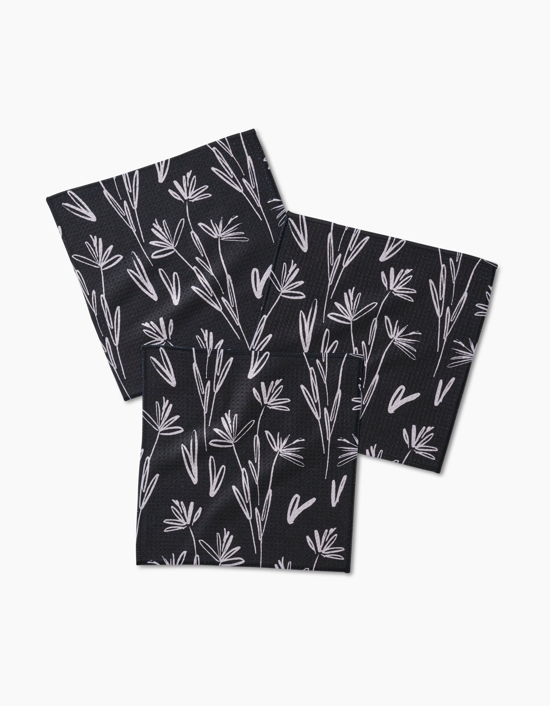 Three “Sketchbook Dusk” napkins by Toboggan Avenue, made of Geoweave fabric with a white floral and leaf design, are arranged overlapping and slightly fanned on a white background.