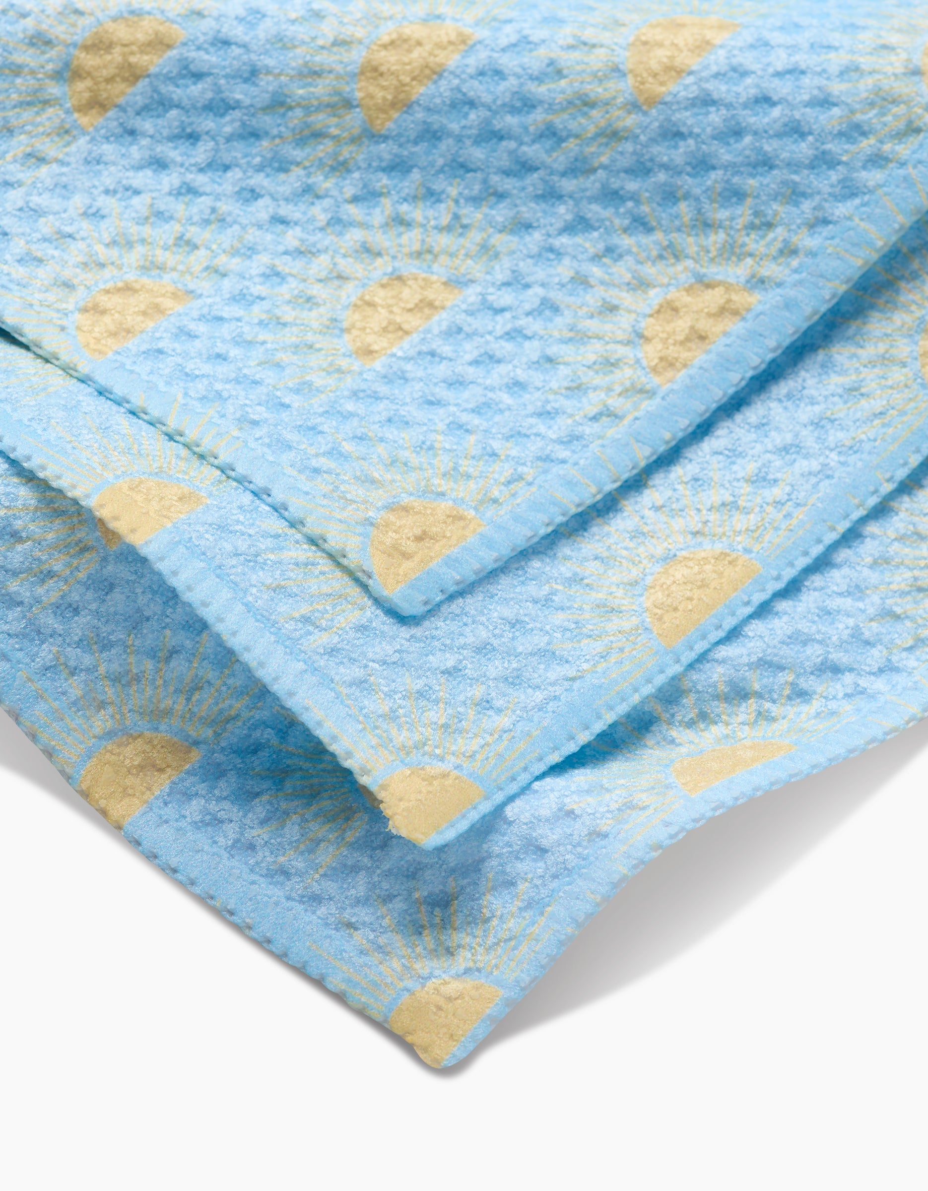 A close-up of GEOMETRY’s Soft Summer Sun towel, made from super-absorbent Geoweave fabric, shows its light blue color, yellow sun pattern, eco-friendly softness, and vibrant design while partially folded.