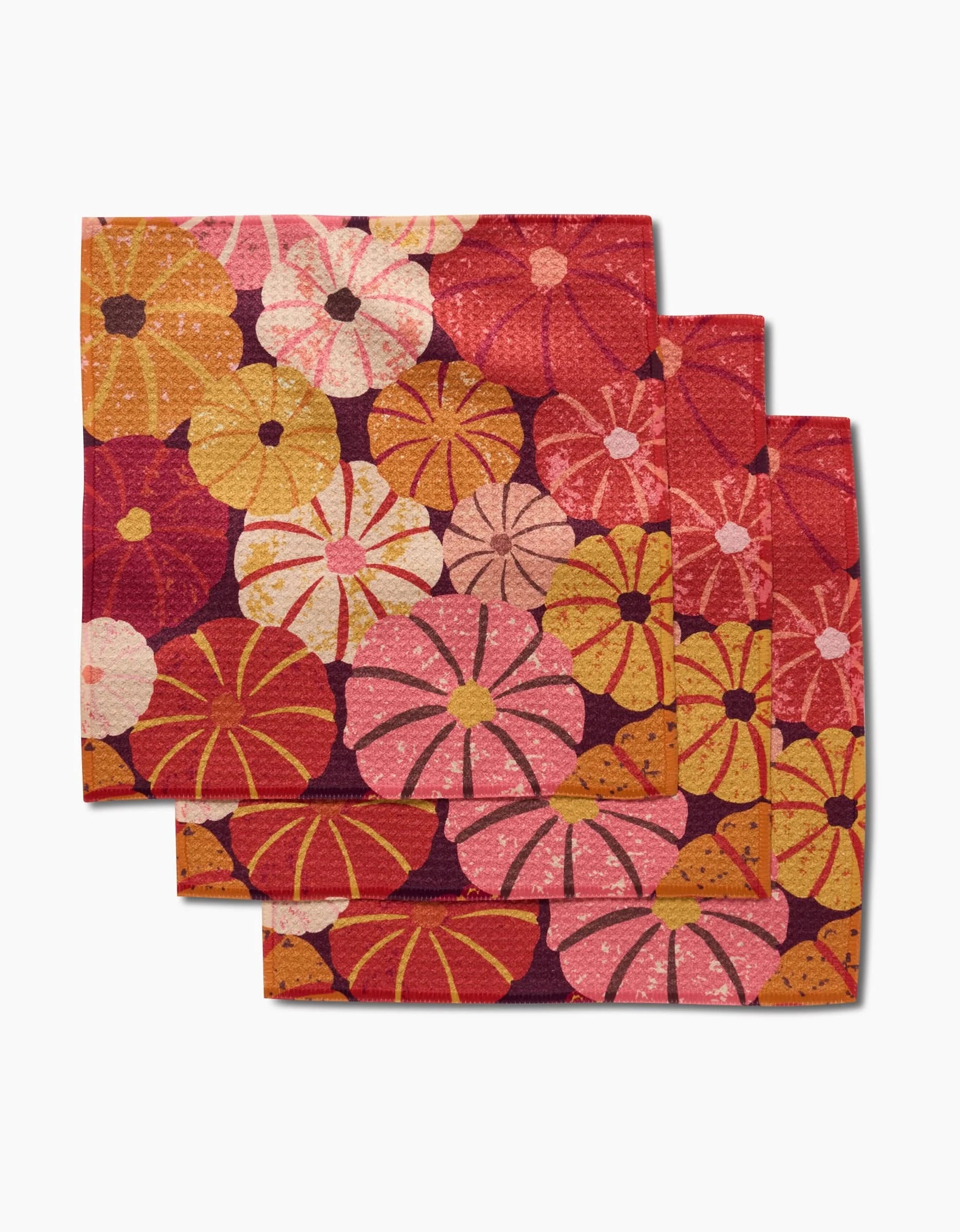 Three square napkins from Rebecca Bobko’s Spicy Pumpkins collection, featuring a vibrant floral pattern in pink, orange, yellow, and red on white—perfect recycled dishcloths to brighten up your kitchen.