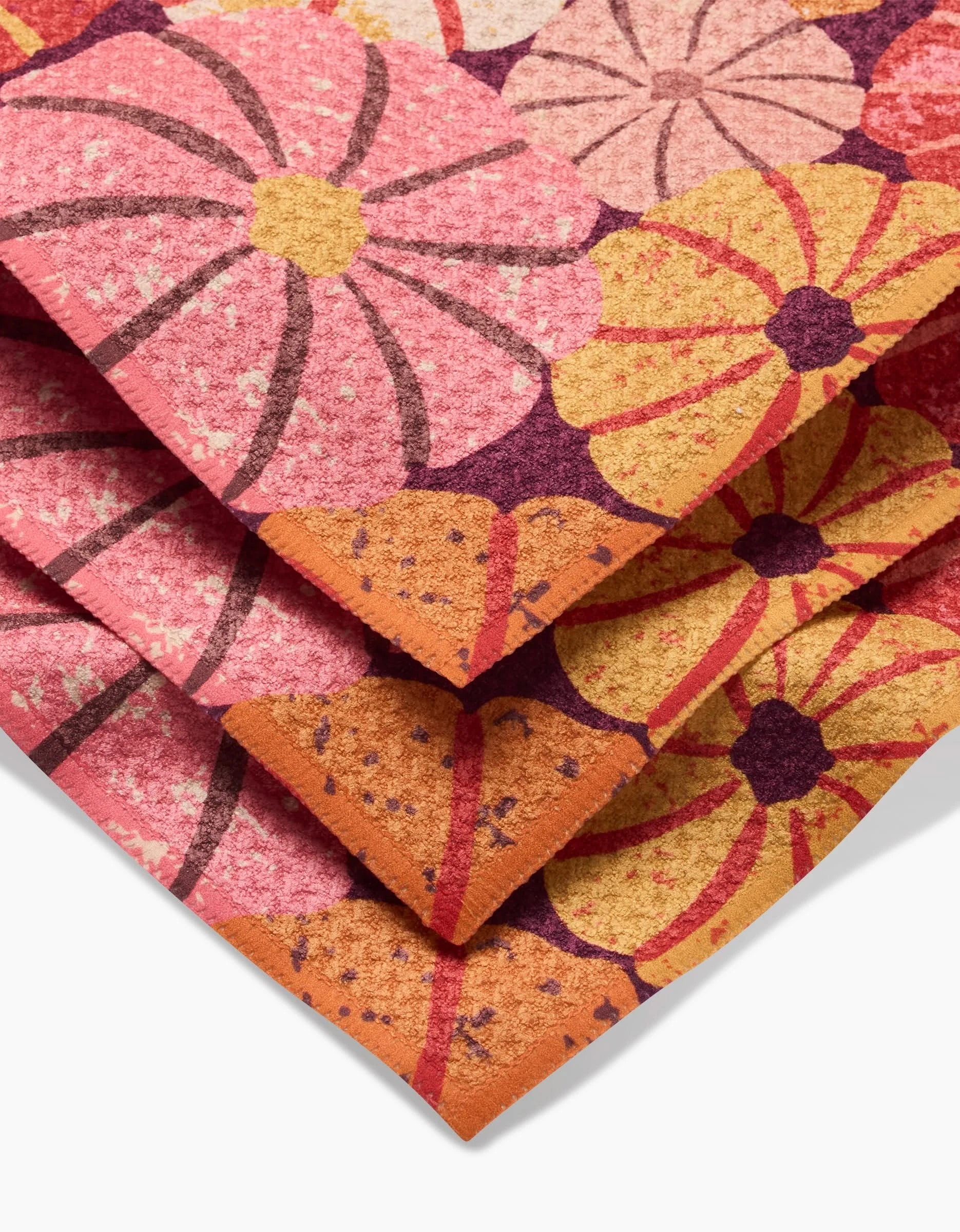 A close-up of the folded Spicy Pumpkins rug by Rebecca Bobko displays large, stylized flowers in pink, orange, yellow, and purple hues with textured details—evoking vibrant recycled dishcloth designs.