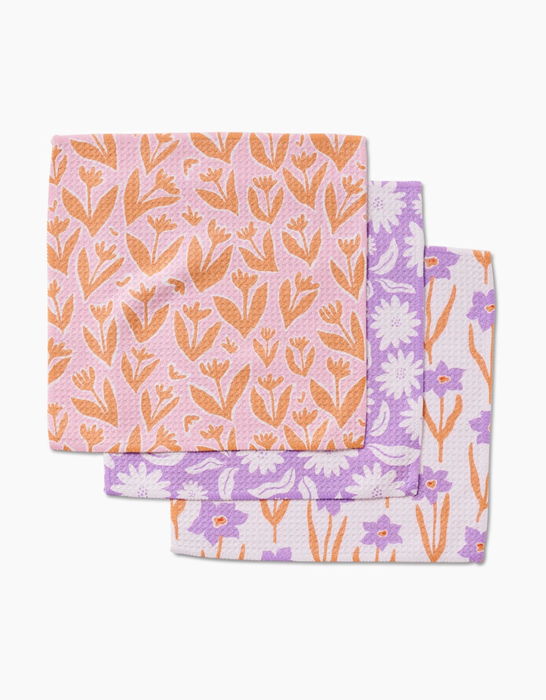Three Spring Flowers dishcloths by Kayla Ann, stacked: orange with peach flowers on top, purple with white flowers in the middle, and white with light purple and orange flowers at the bottom—all made from super-absorbent Geoweave fabric.