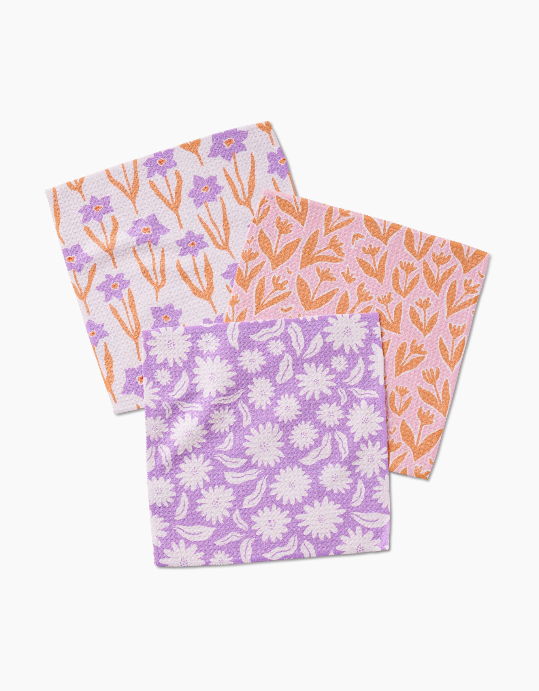 Three Kayla Ann Spring Flowers dishcloths, made from super-absorbent Geoweave fabric, are arranged overlapping: one with purple and white daisies, one with orange leaf motifs, and one with purple flowers and orange stems.