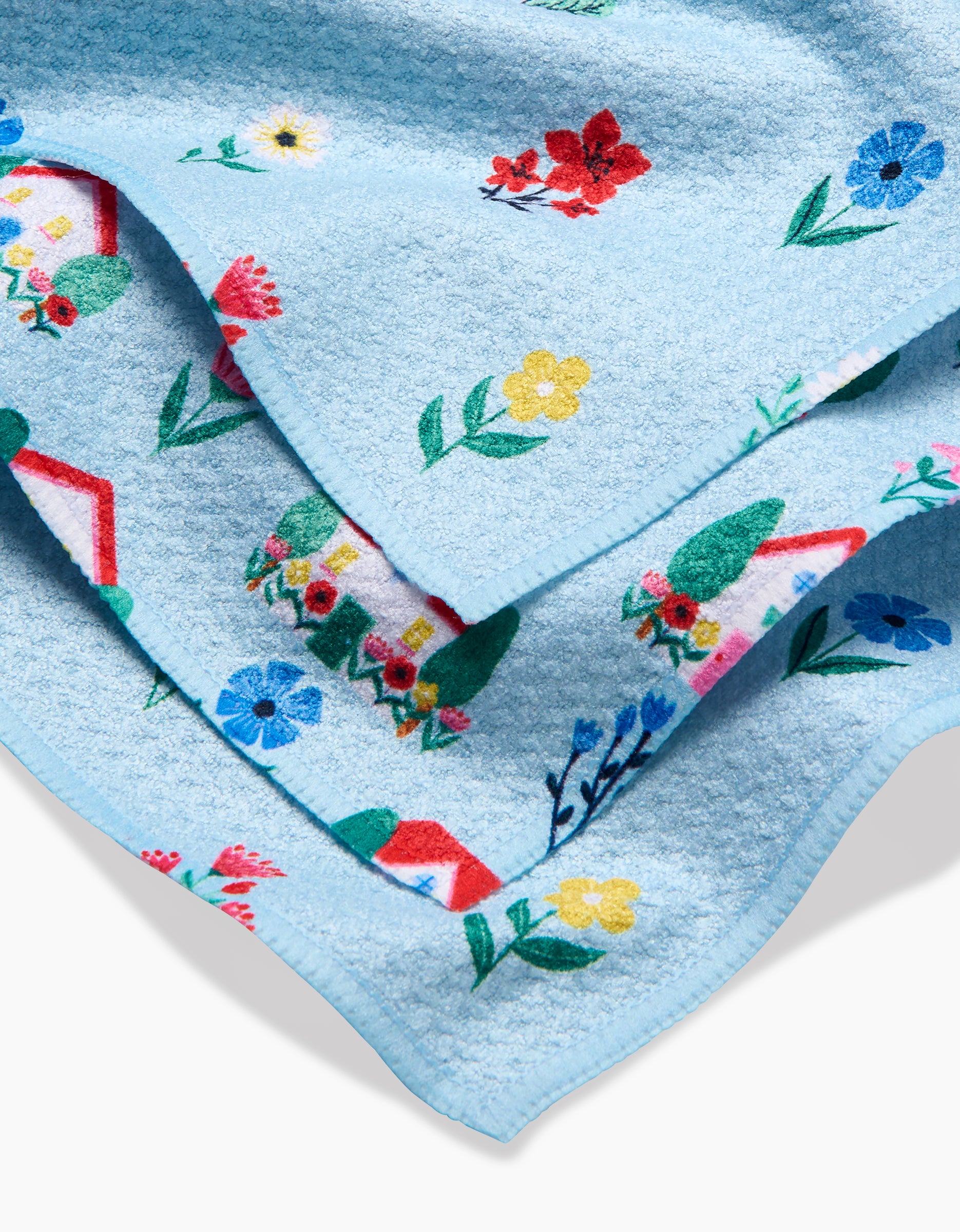 The Spring House Blossom blanket by Ceyda Alasar features light blue textured Geoweave fabric with colorful prints of flowers, houses, and plants in red, yellow, green, and blue, finished with a stitched edge and shown partially folded.