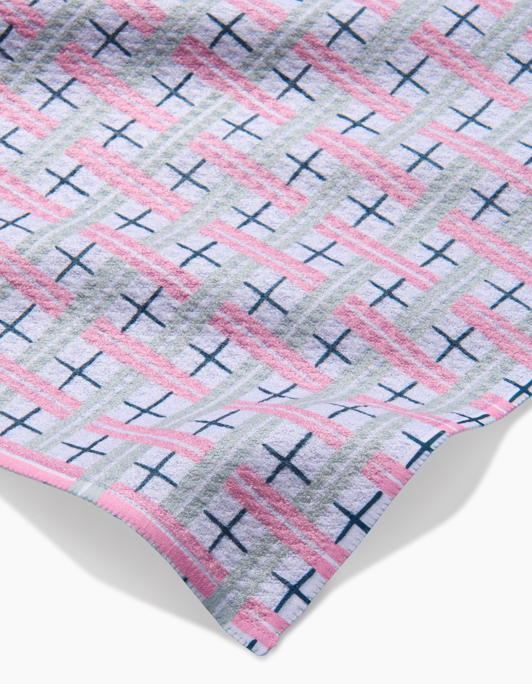 A close-up of Hufton Studio’s Spring Picnic Day dishtowel shows its super-absorbent Geoweave fabric with a geometric pattern of pink, gray, and blue intersecting lines forming a grid with blue crosses and a slightly wrinkled edge.