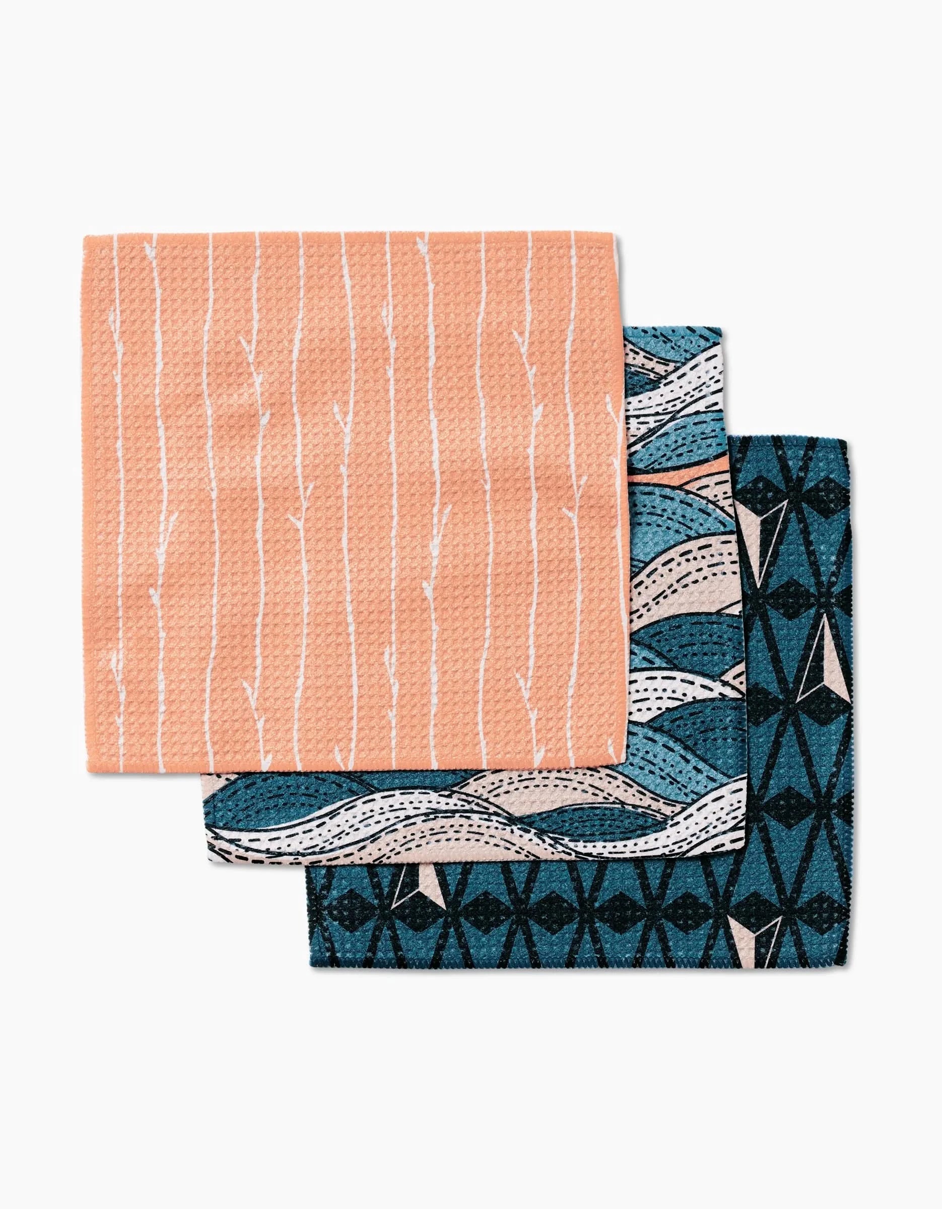 Three folded Twig + Spark Squall dishcloths are stacked: the top is peach with white branches, the middle has blue and white abstract waves, and the bottom is dark blue with a geometric black and white design. Made from Geoweave fabric.