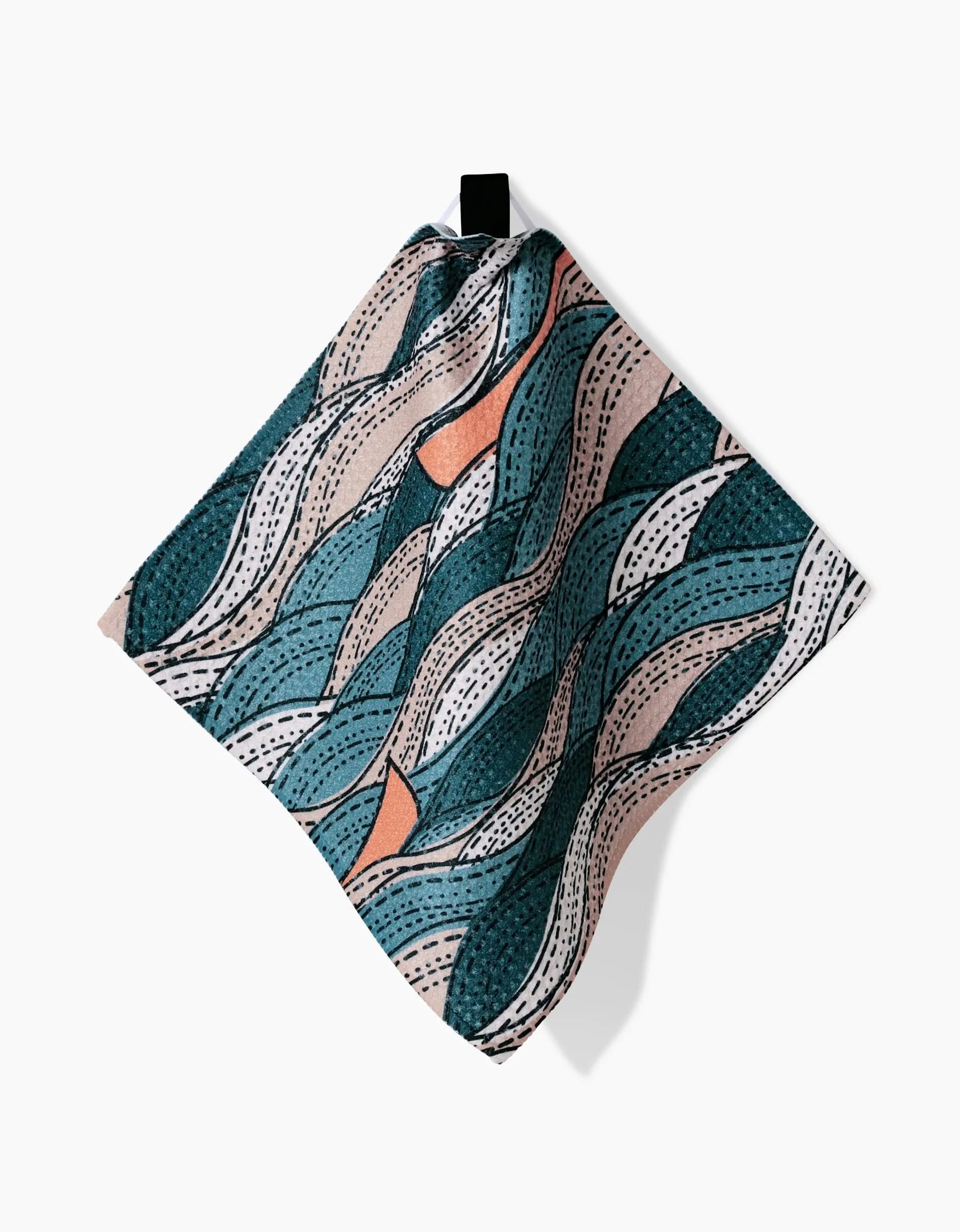 The Twig + Spark Squall dishcloth, made from Geoweave fabric, displays wavy teal, peach, and beige shapes with dotted lines, and hangs from a black loop against a white background.