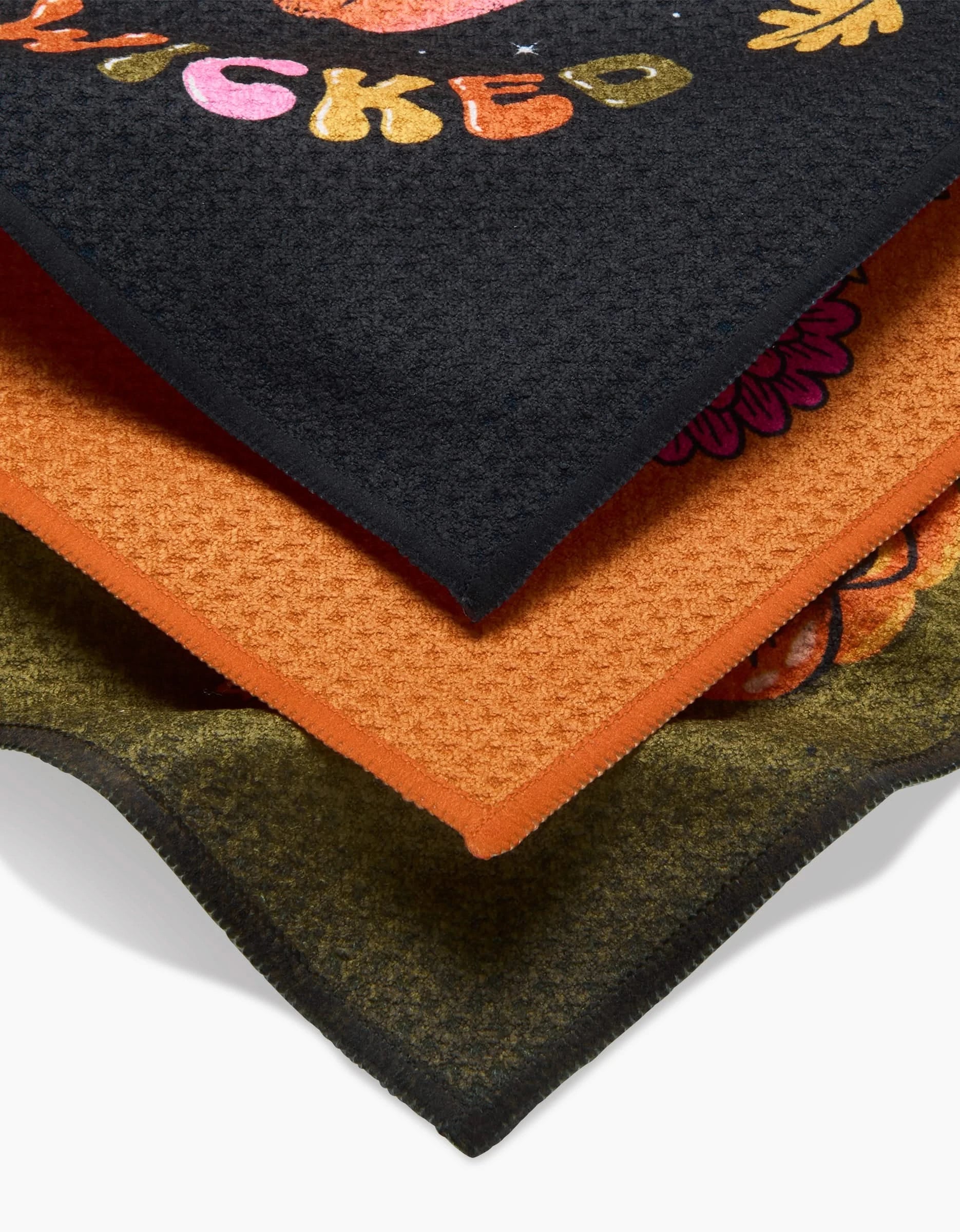 The Stay Weird Stay Wicked dishcloth set by Megan Roy includes three super-absorbent towels: a black towel with playful lettering and florals, an orange solid towel, and an olive green towel with a partial orange pattern.