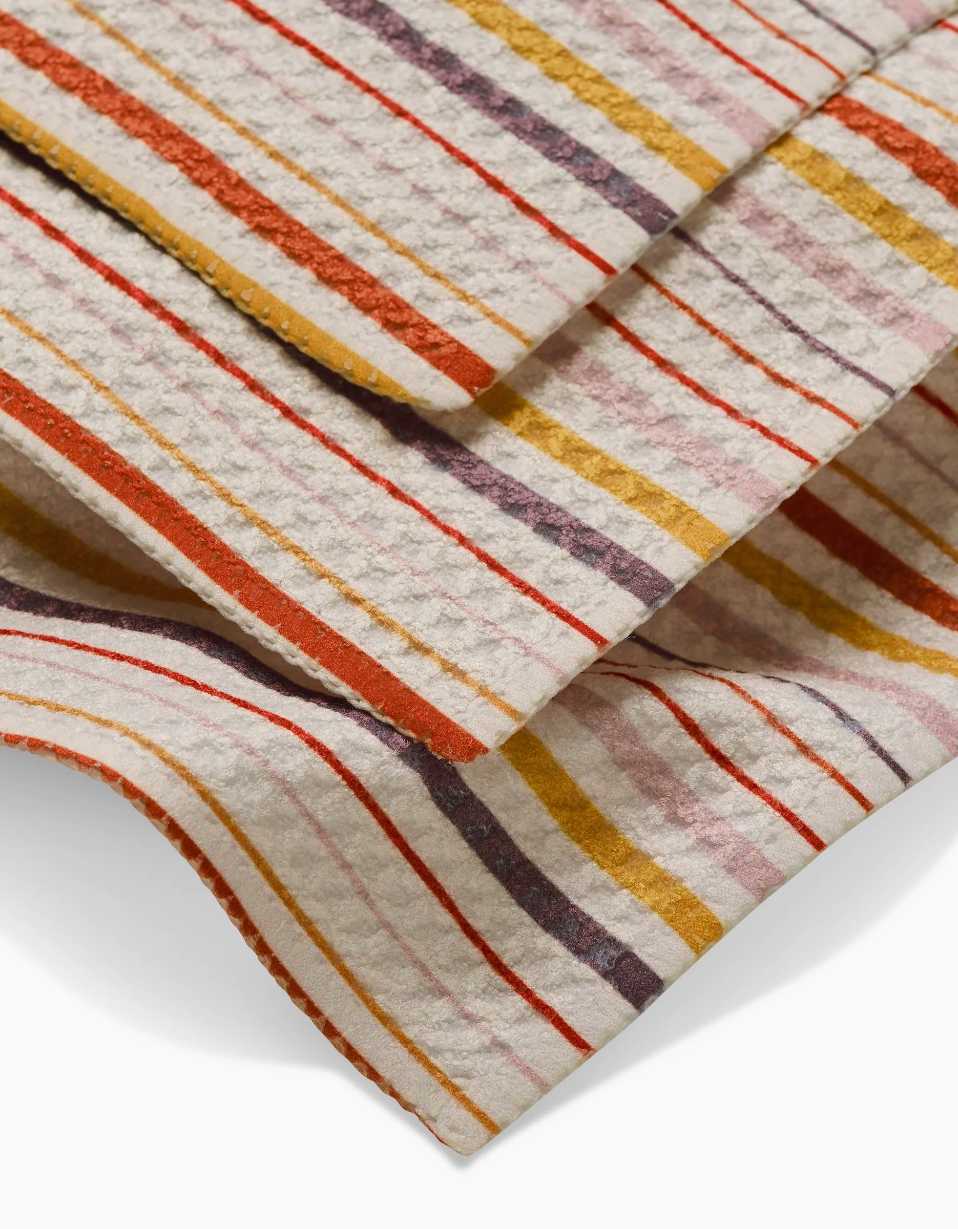 Close-up of GEOMETRYs Stripe Season dishcloth fabric showing textured, super-absorbent material with horizontal stripes in red, orange, yellow, purple, and beige on an off-white background; edges appear folded.