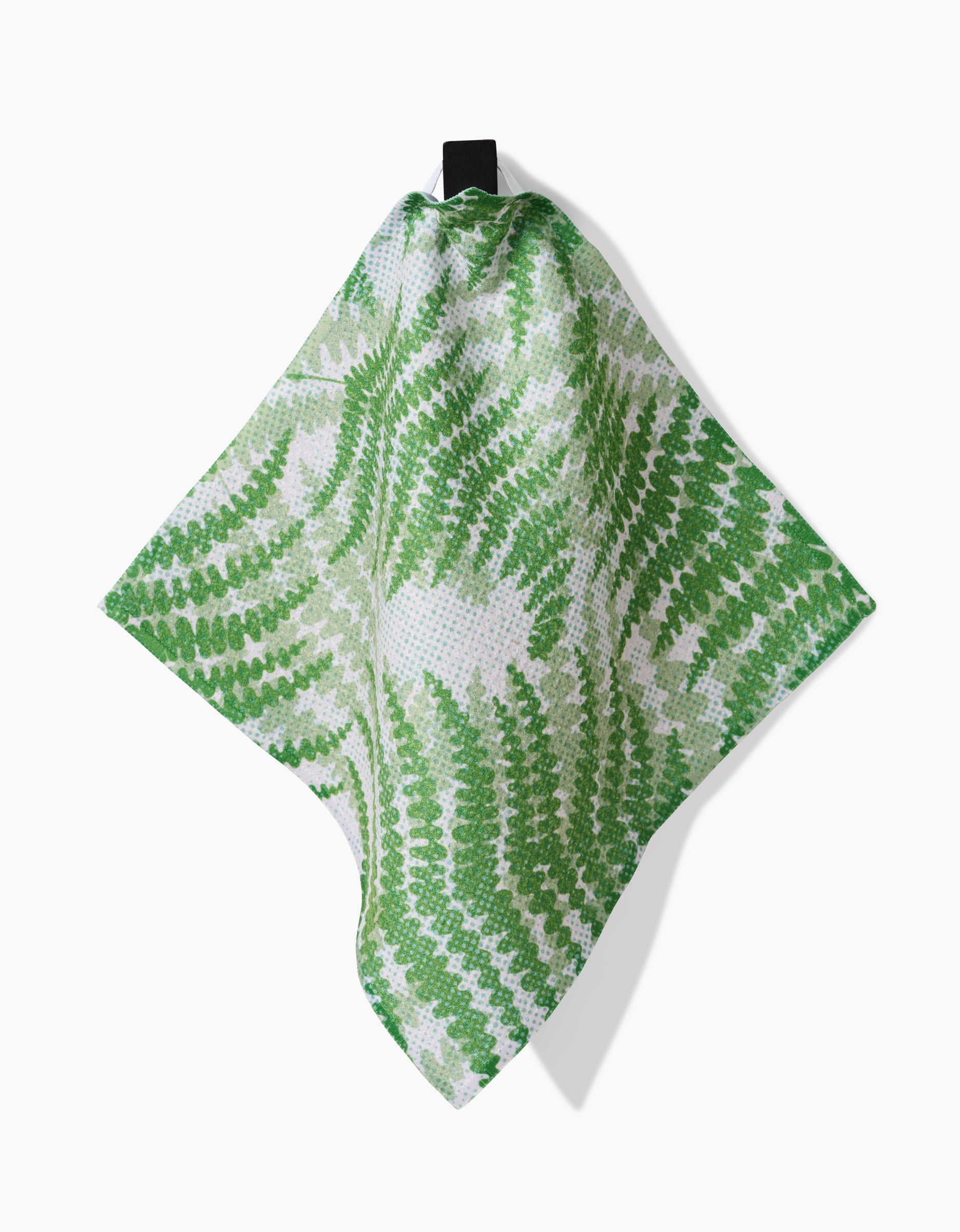 The GEOMETRY Swaying Leaf dishcloth, crafted from recycled materials, features a green and white fern-inspired pattern and hangs on a black hook against a white background—combining style with sustainability.