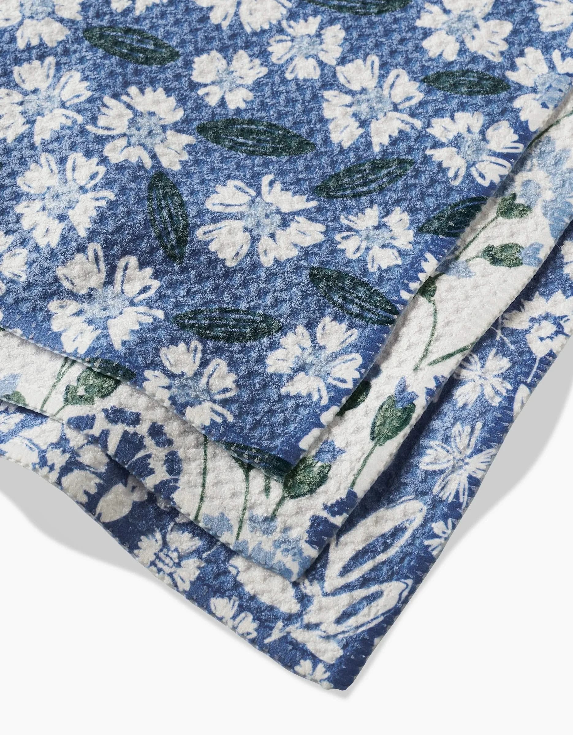 Three folded blue Sweetie Floral Geoweave dishcloths by Hufton Studio, each with unique white floral and green leaf patterns, are stacked against a white background. Eco-friendly and stylish, they bring charm to any kitchen.