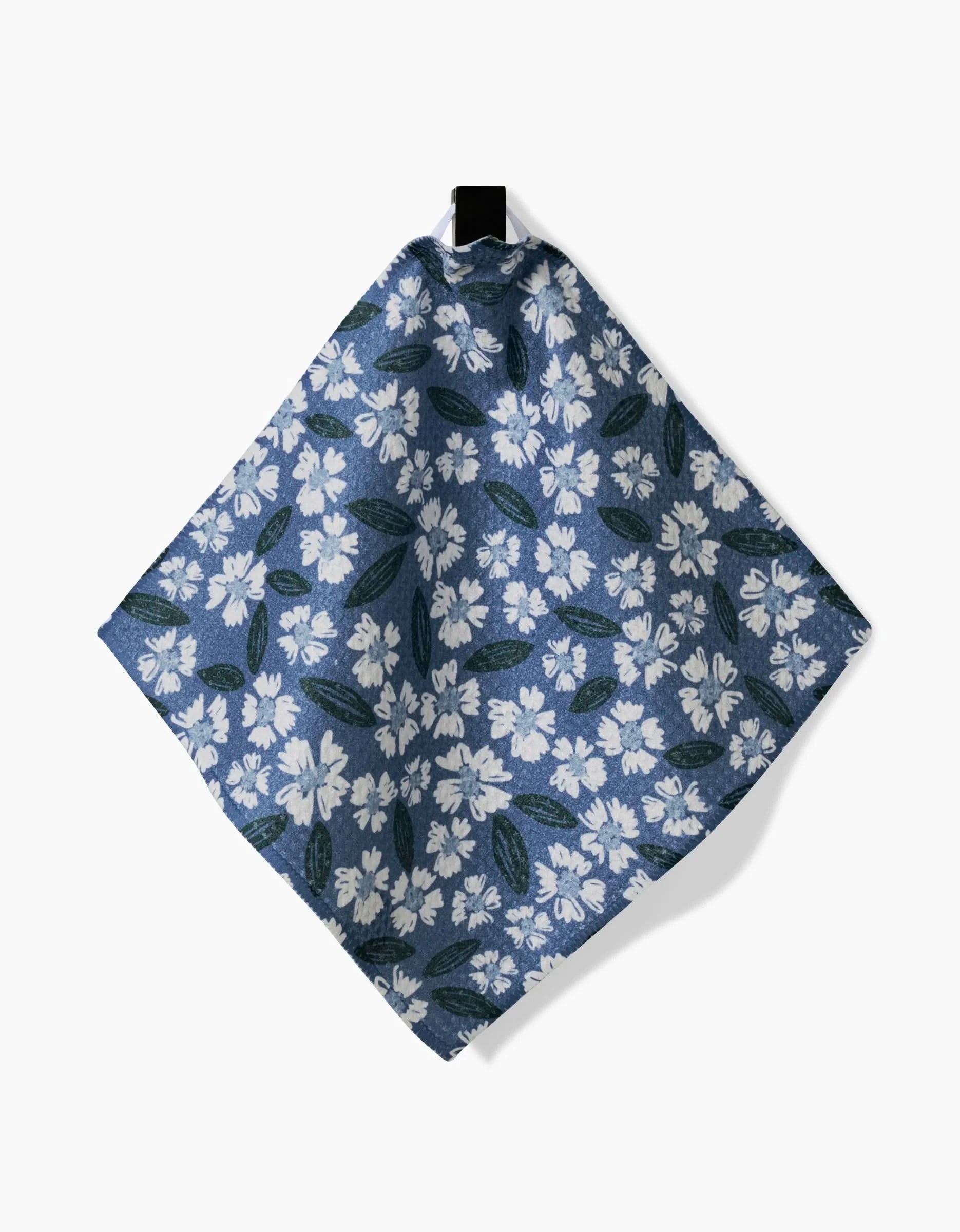 The Hufton Studio Sweetie Floral Geoweave dishcloth features a blue background with white flowers and green leaves, hanging from a black hook—an eco-friendly kitchen accessory.