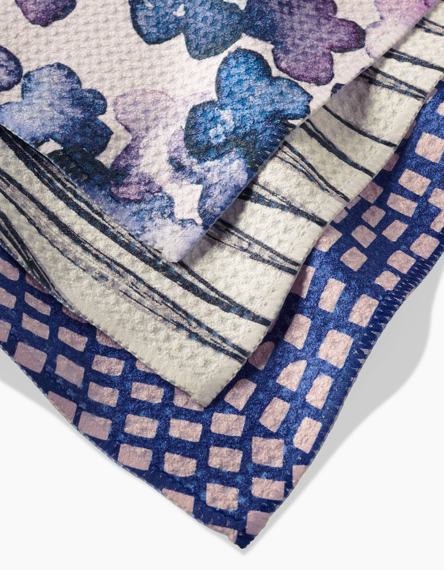 Three overlapping GEOMETRY Tiny Petals dishcloths: one with blue and purple watercolor flowers, one with navy and white stripes, and one featuring a blue and pink geometric grid design, all crafted from Geoweave fabric.