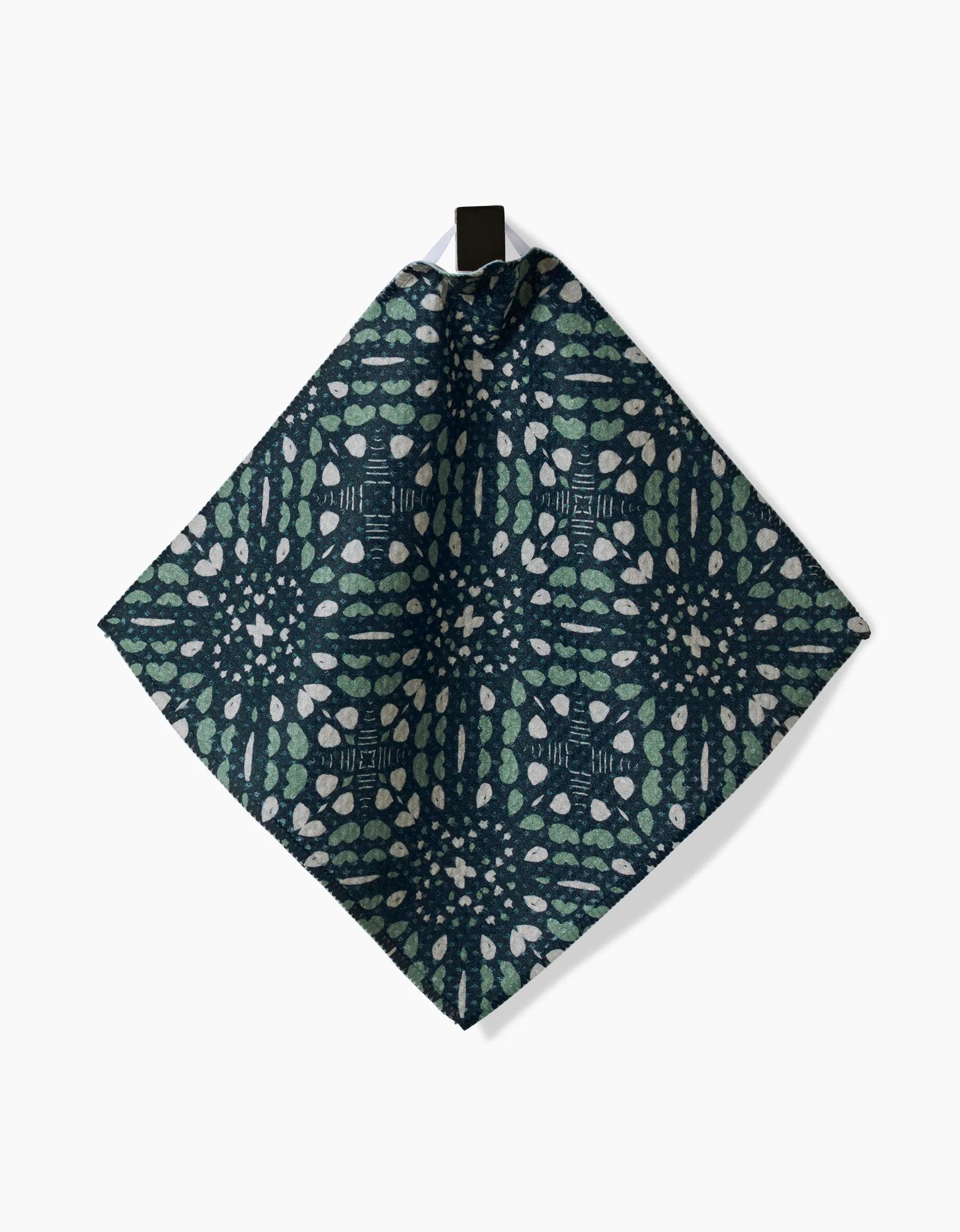 The Twig + Spark Trail Dusting napkin features a diamond shape in dark blue Geoweave fabric with a green and white geometric pattern, hanging from a black loop against a white background.