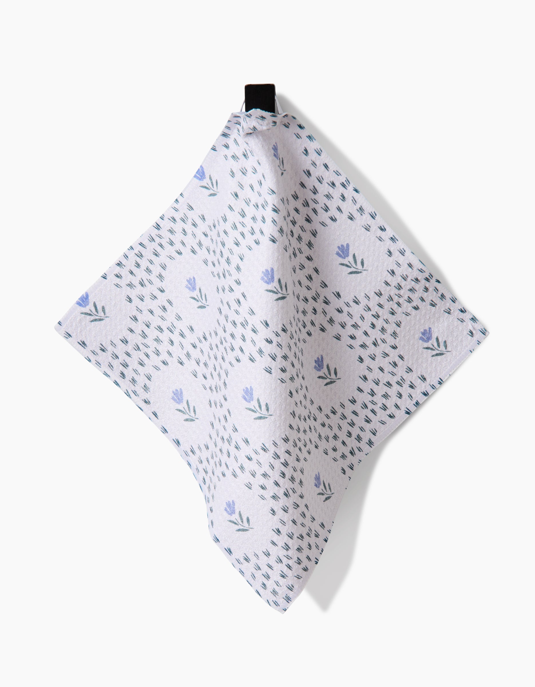 A Trailing Farmhouse Florals napkin by Teri the Butter, made from light-colored Geoweave fabric and featuring small blue flowers with green leaves, hangs from a black loop against a white background.