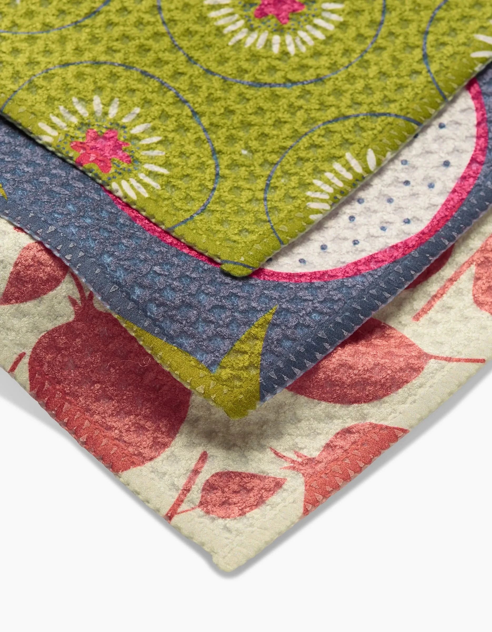 Three textured fabric pieces from the Lizzie Clark Tropical Fruit Dishtowel Set feature colorful patterns: green with circular floral designs, blue with polka dots and pink accents, and cream with red fruit-like motifs.
