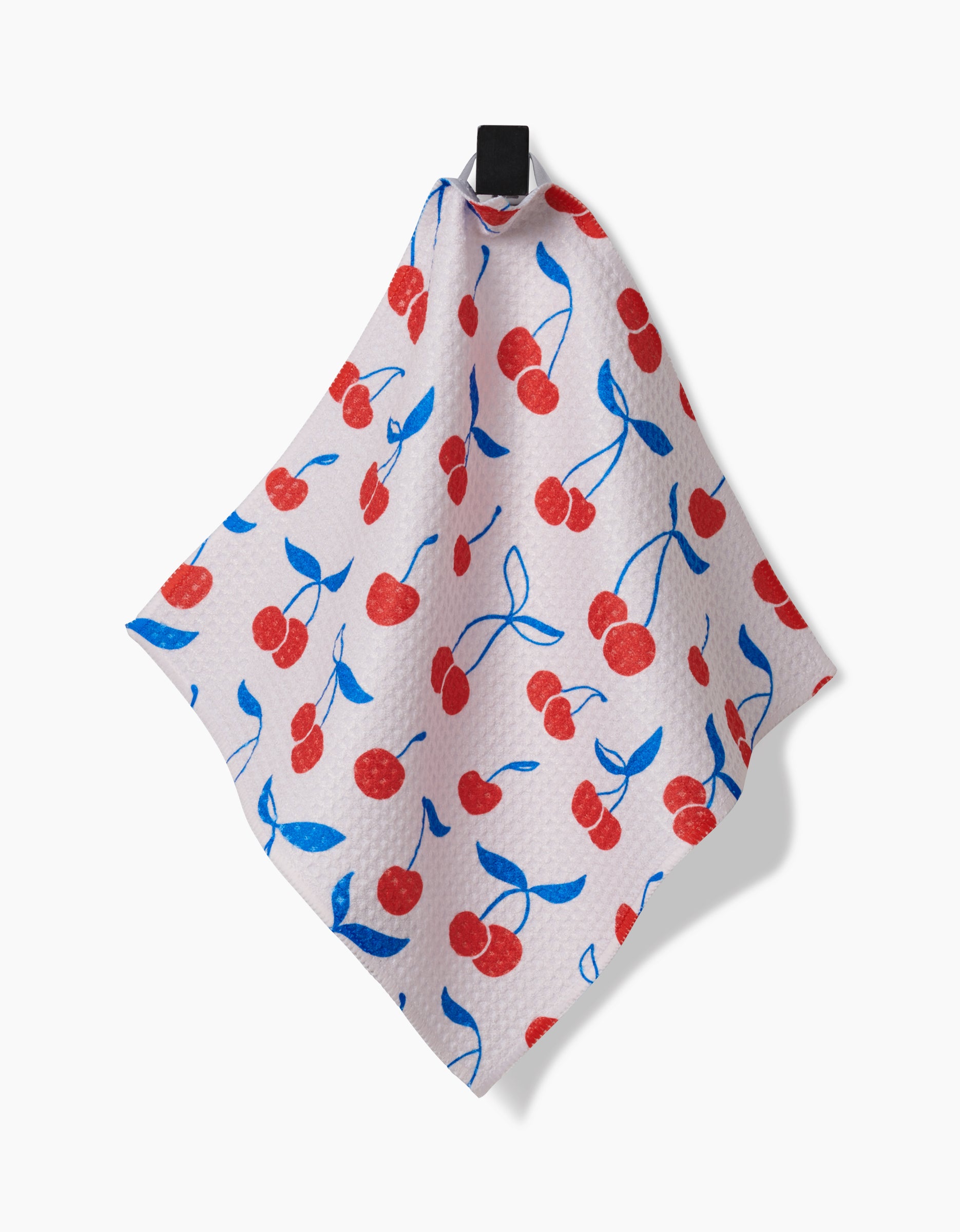 The Kayla Ann Very Cherry kitchen towel, made from super-absorbent Geoweave fabric and adorned with red cherries and blue leaves, hangs from a black hook against a plain white background.