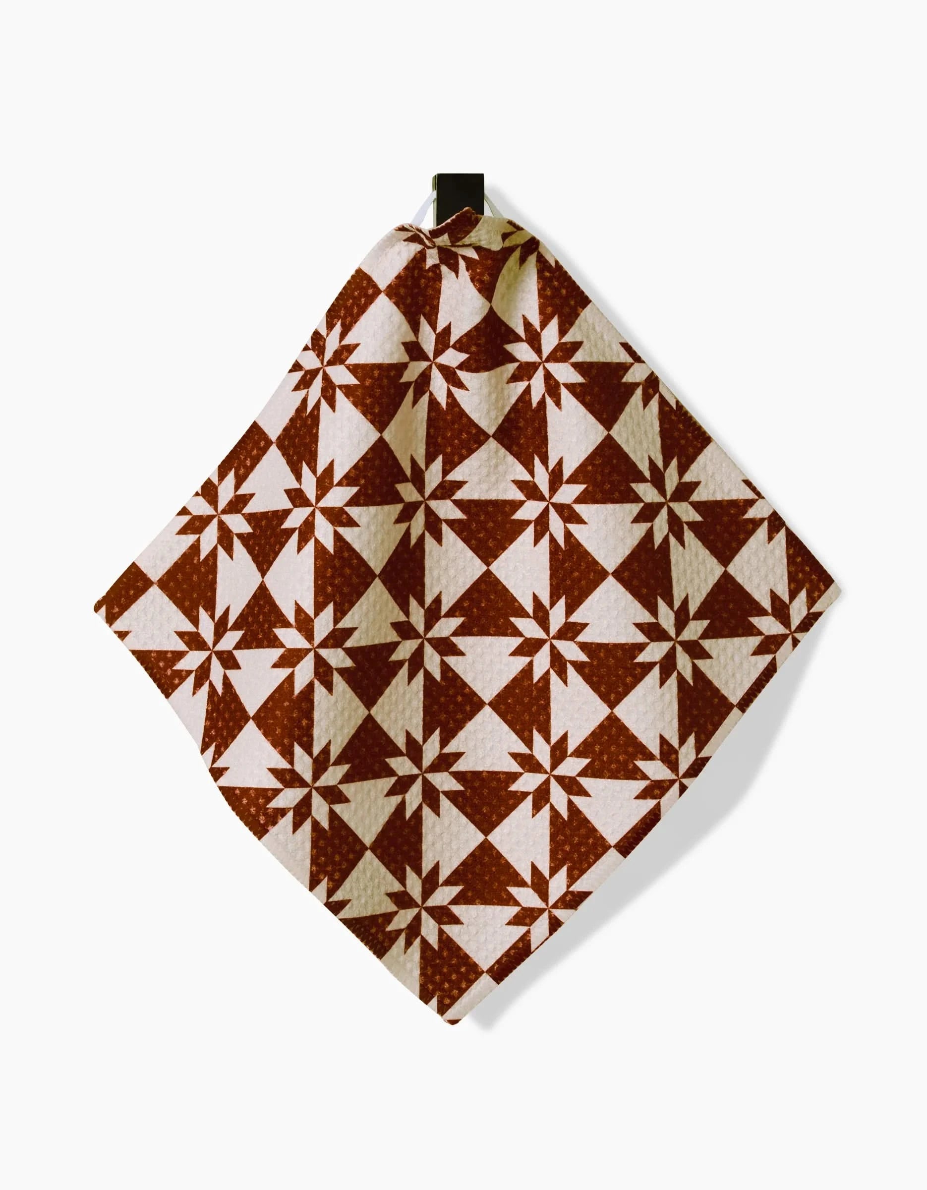 Aubrey Fairchilds Vintage Christmas Geoweave dishcloth, made from recycled materials and featuring a repeating geometric star and triangle design in brown and white, hangs from a black hook against a plain white background.