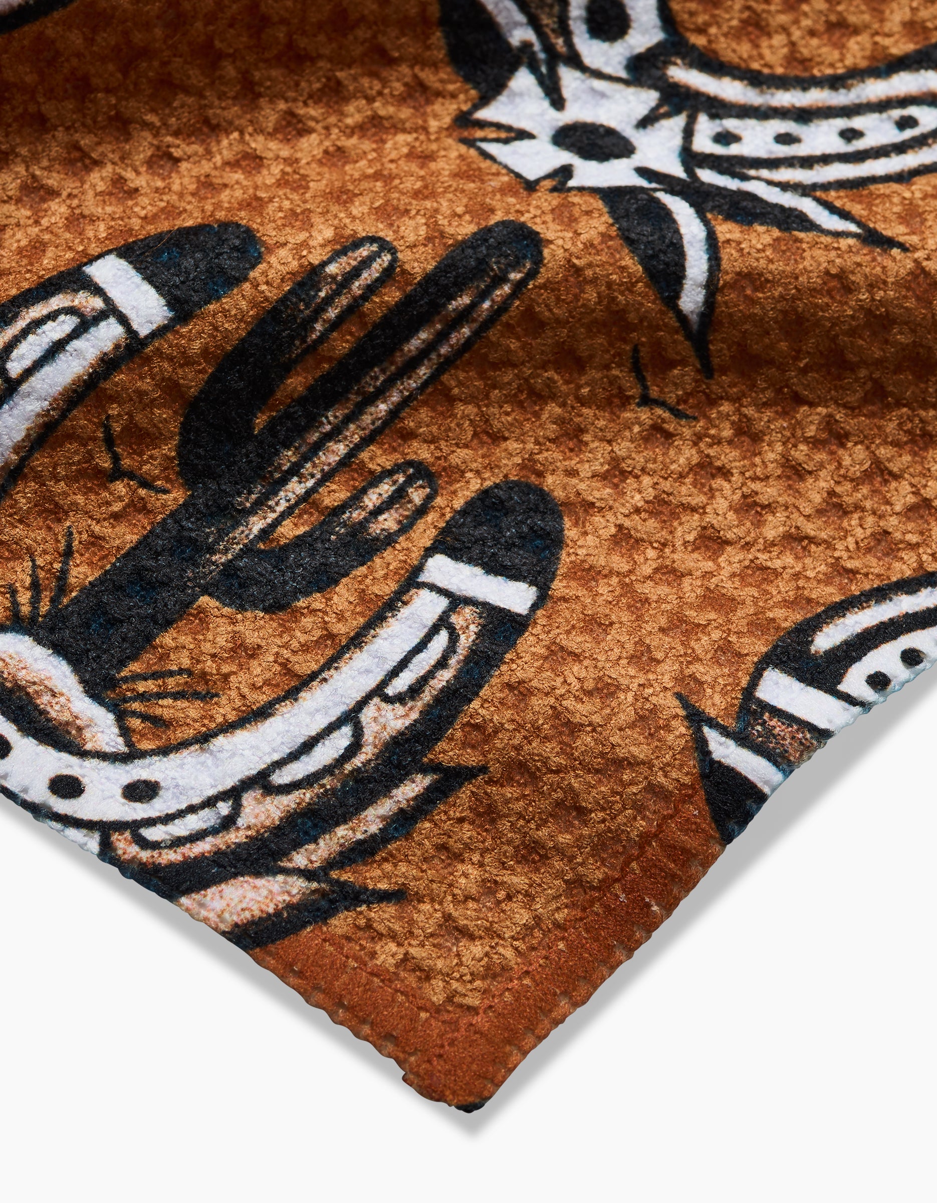 Close-up of Cole Strems Western Horseshoes brown Geoweave dishcloth, featuring black-and-white cacti and horseshoe illustrations with a stitched edge visible in the lower right corner.