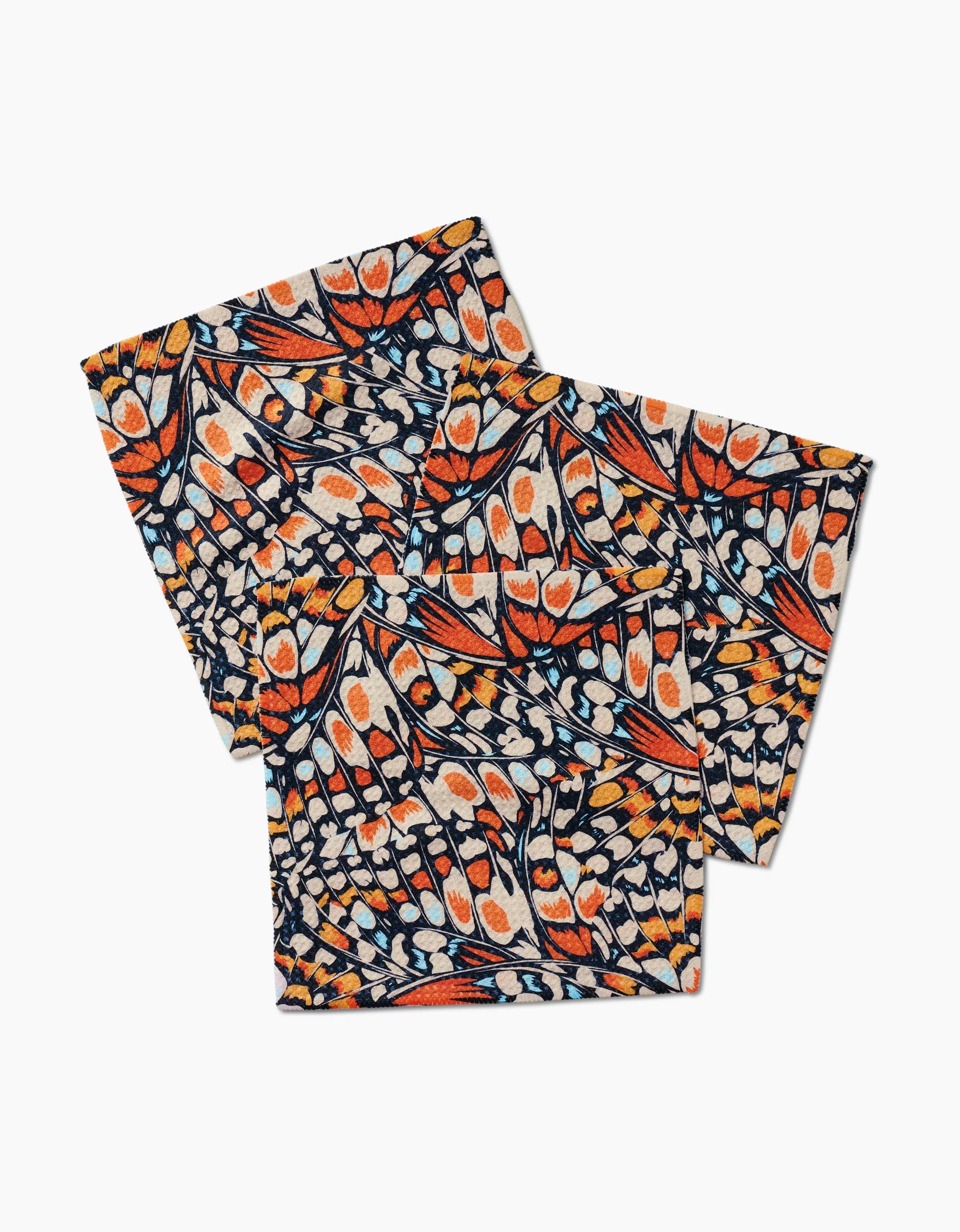 Two folded GEOMETRY Wings in the Wind dishcloths, made of Geoweave fabric, display a vibrant butterfly wing pattern in orange, black, white, and blue on a light background. They are overlapping on a white surface.