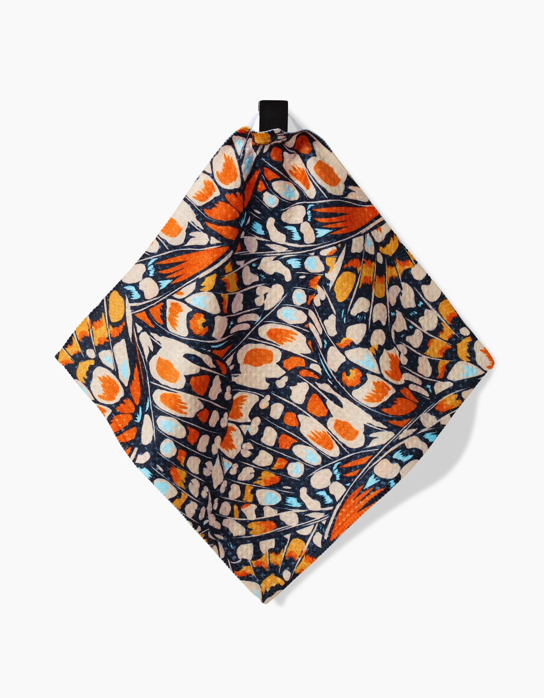 The GEOMETRY Wings in the Wind napkin is a square, recycled fabric with a vibrant butterfly wing pattern in orange, blue, black, and beige, shown hanging by a black loop against a white background.