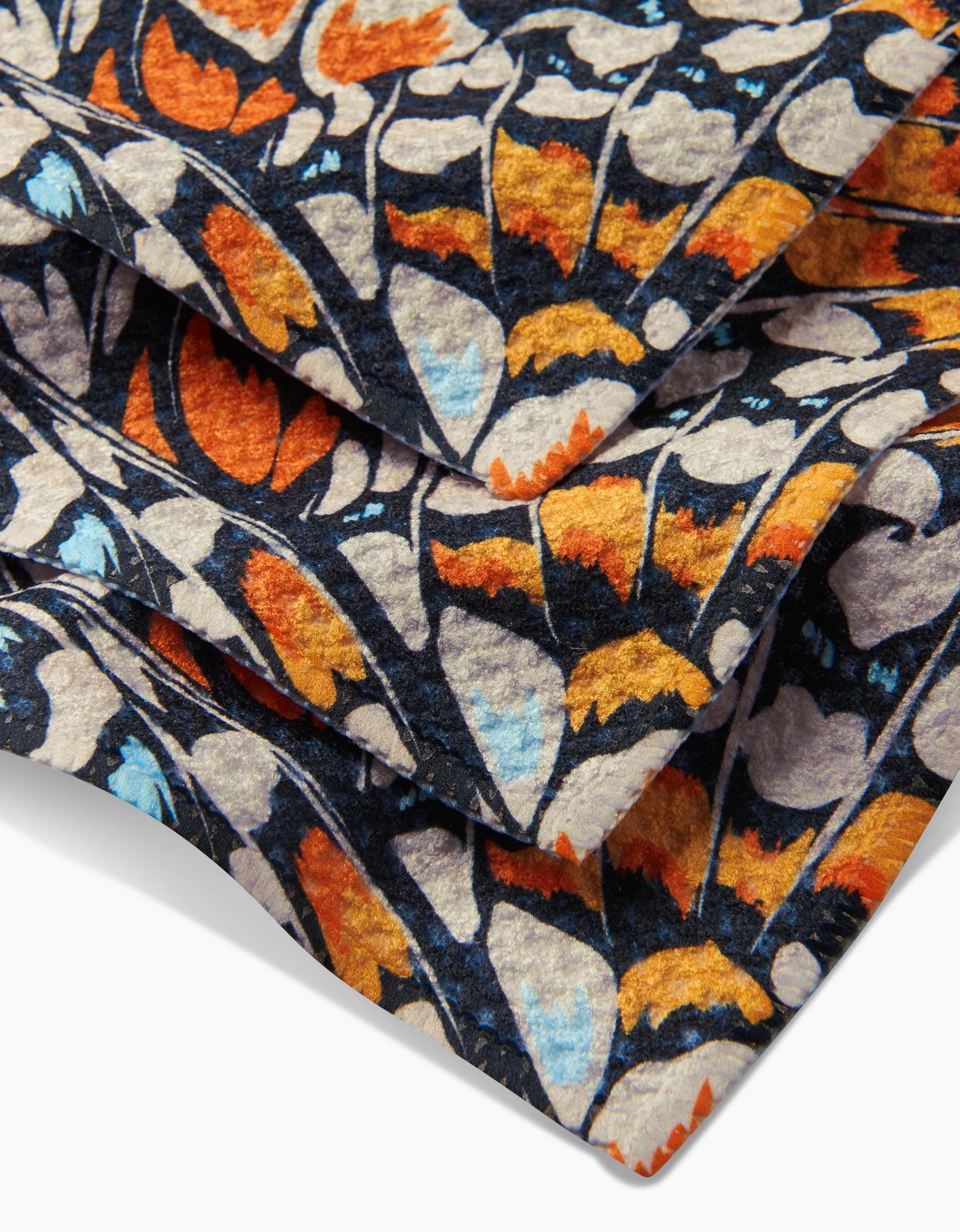 A close-up of GEOMETRYs Wings in the Wind dishcloths, showcasing colorful orange, yellow, light blue, and white floral patterns on a dark geoweave fabric background.