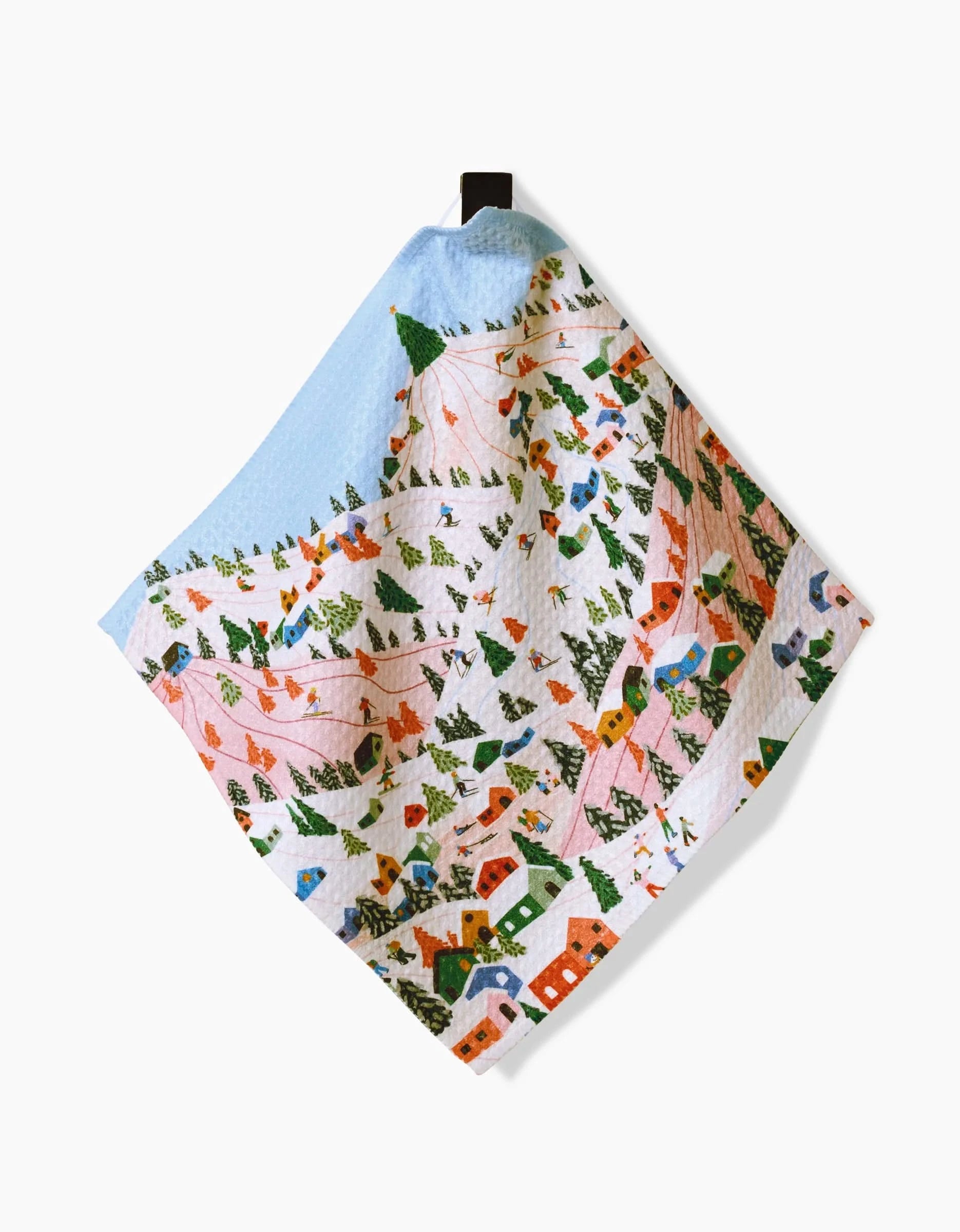 The Winter Holiday dish towel by Ceyda Alasar is made from recycled materials and features a winter village with snow-covered houses, pine trees, and skiers on pink slopes. It hangs by a black loop and is displayed against a white background.