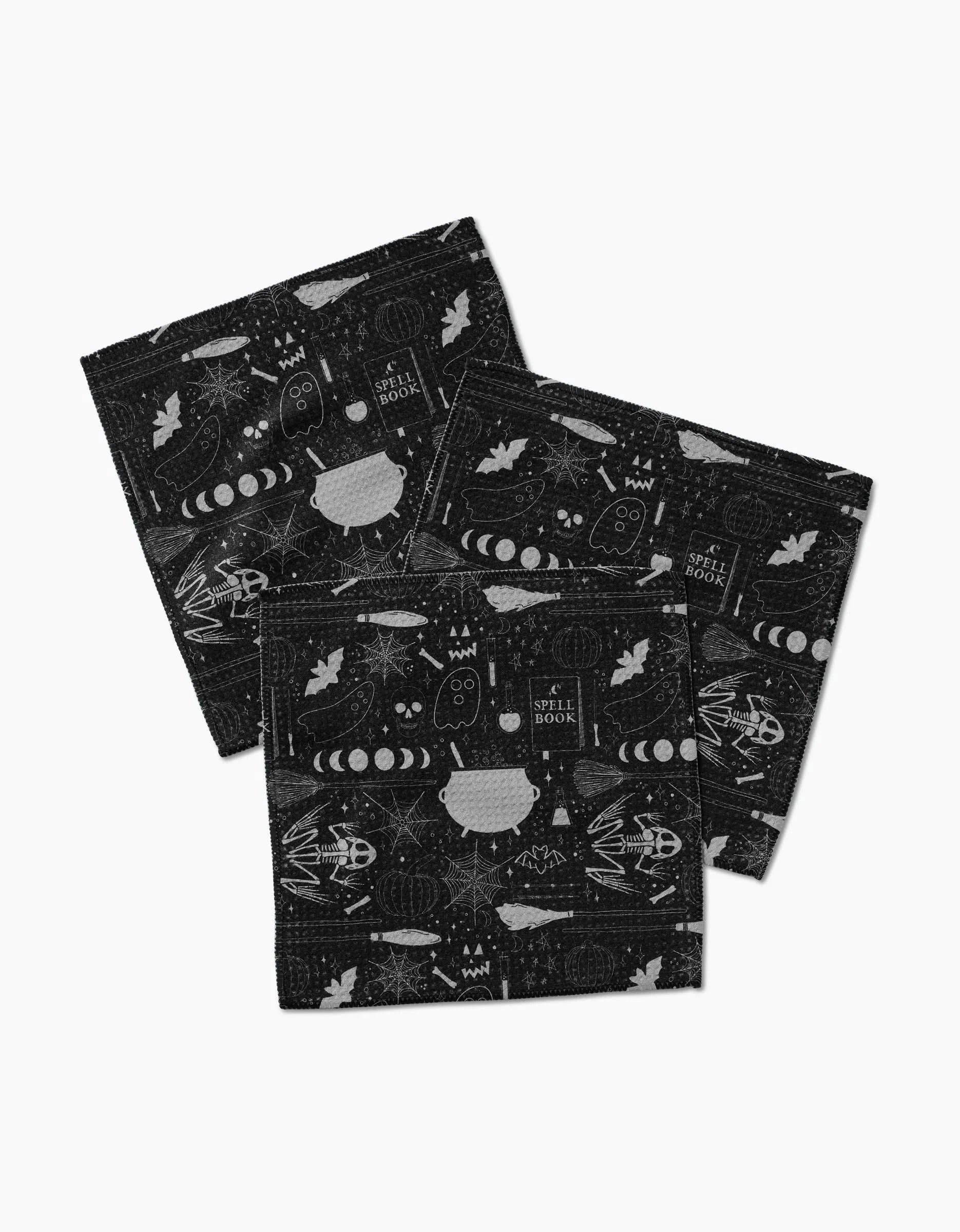 The Witches Brew napkins by Rosa Kichline include three black cloths made from absorbent Geoweave fabric, each showcasing white illustrations of cauldrons, spell books, moons, owls, leaves, insects, and stars.
