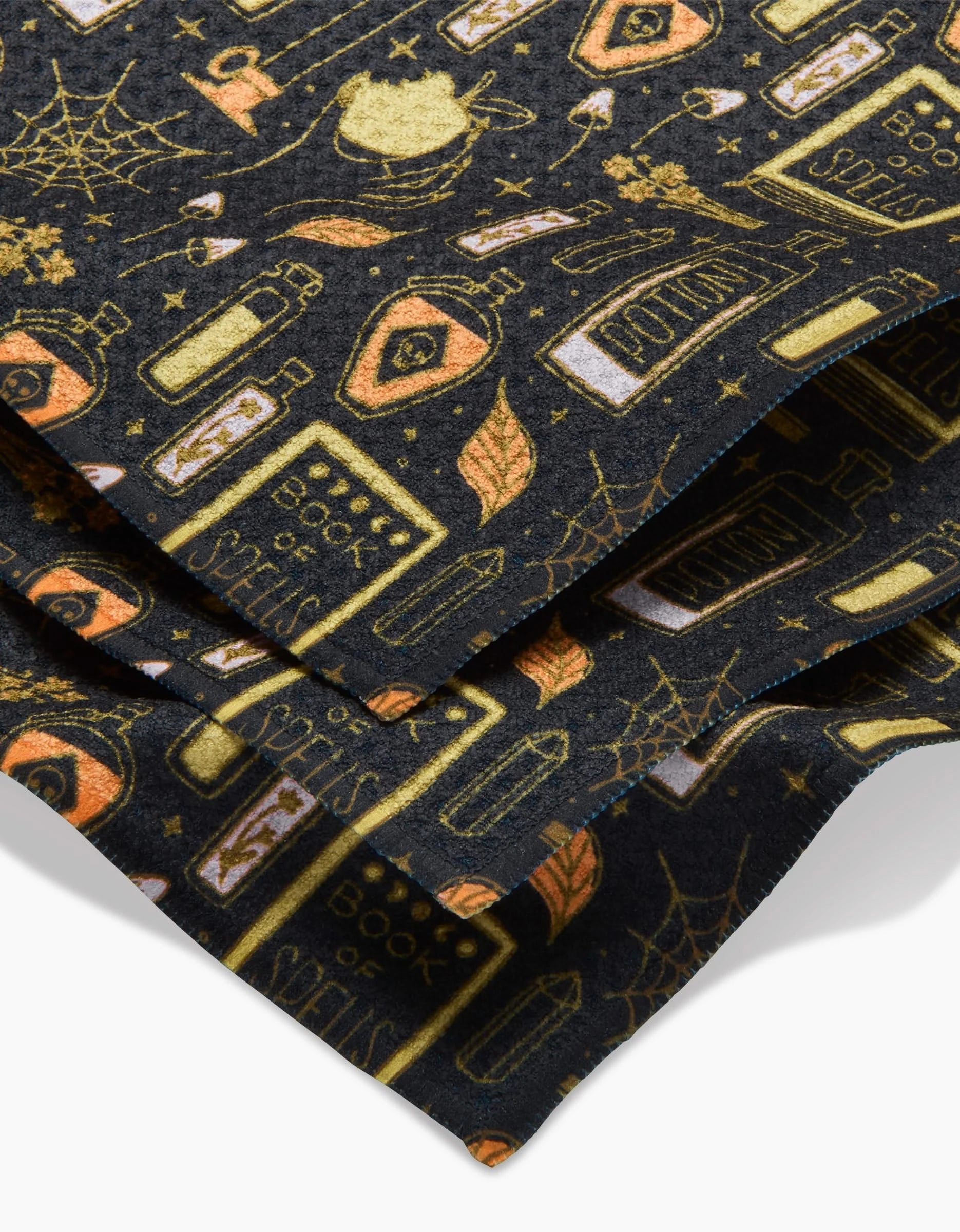 The Witchy Things dishtowel set by Mandy Arroyo features black fabric with a Halloween pattern—potion bottles, Book of Spells, spider webs, and crystals in gold and orange. Made from recycled materials, these super-absorbent towels are neatly folded.