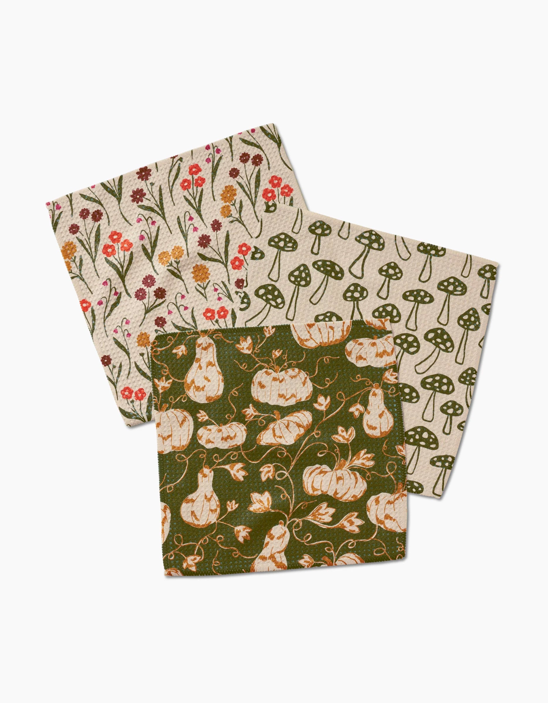 Kayla Anns Woodland Trail set includes three dishcloths—red and orange wildflowers on cream, green mushrooms on beige, and white pumpkins with vines on green—artfully overlapped against a white background.