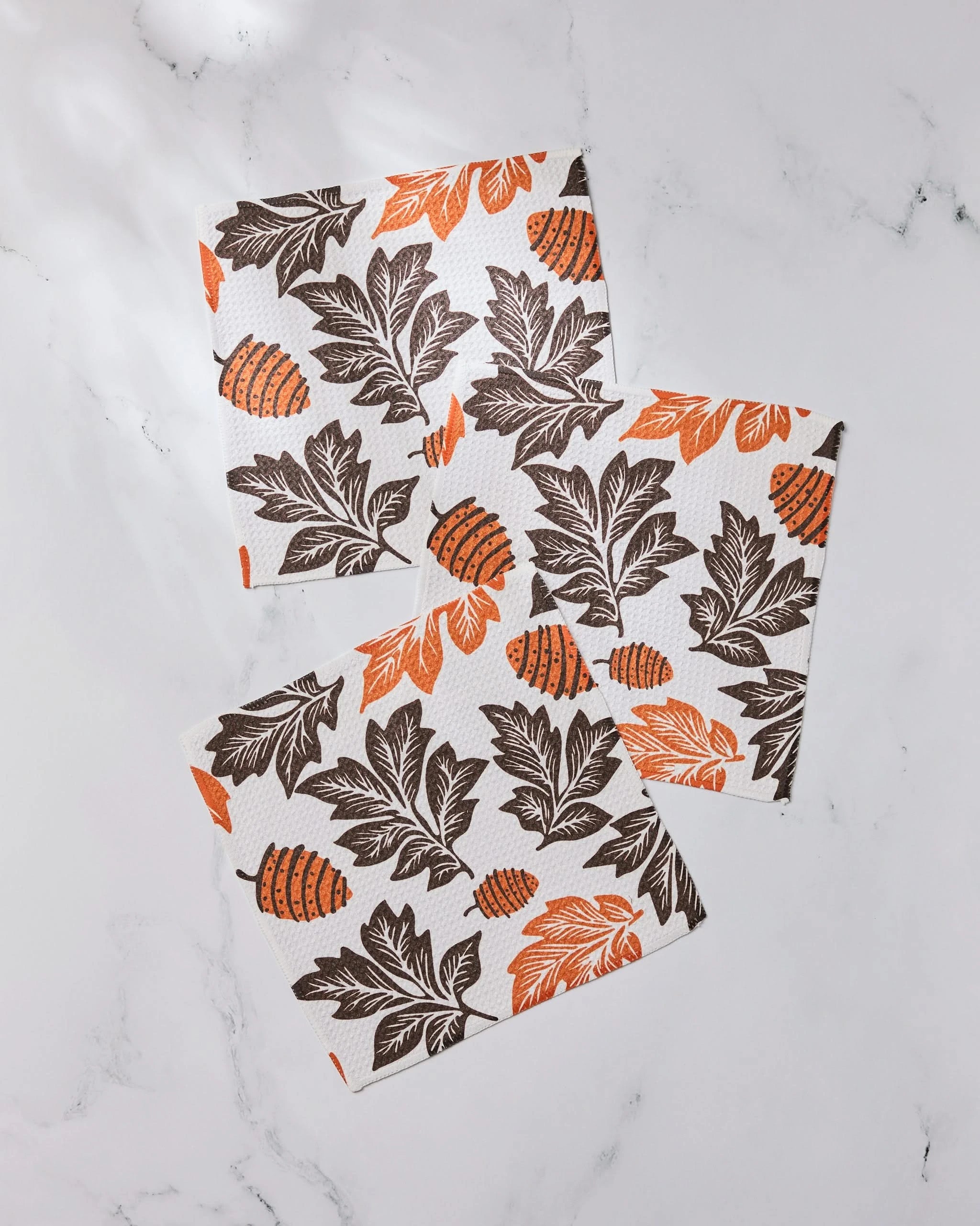 The Geometry Woodland Waltz set includes four napkins made from recycled materials, featuring a fall-themed pattern of brown leaves and orange pinecones on a white marble background.