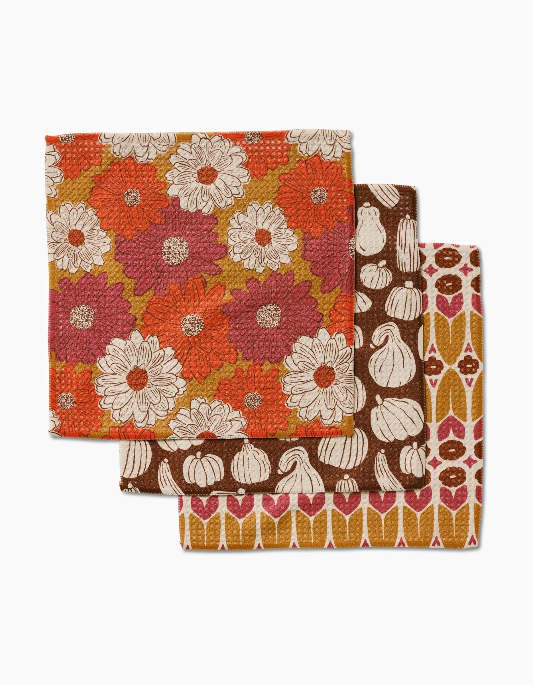 The Kayla Ann Zinnia Forever set includes three folded dishcloths made from recycled materials, featuring fall-themed patterns: orange, red, and white flowers; white gourds on brown; and orange, pink, and white geometric shapes.