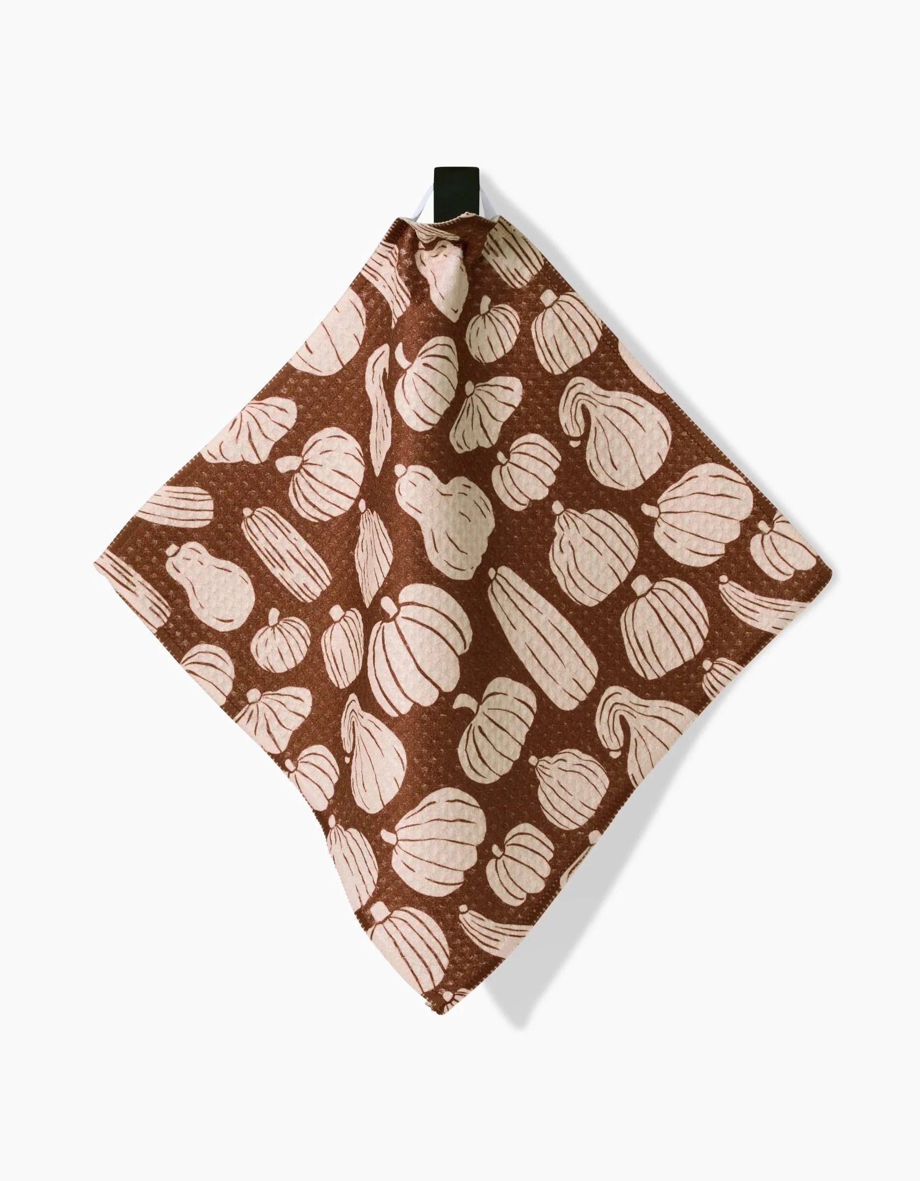The Kayla Ann Zinnia Forever is a brown recycled cloth towel with light gourd and pumpkin patterns, hanging from a black loop against a white background.