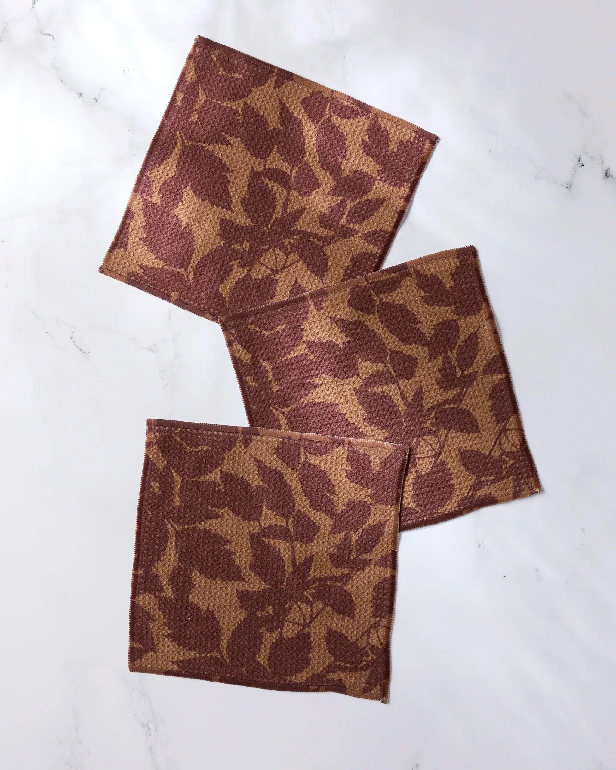 Three Rustle square cloth napkins by Geometry, featuring a brown and tan leaf pattern and made from recycled materials, are arranged overlapping on a white marble surface.