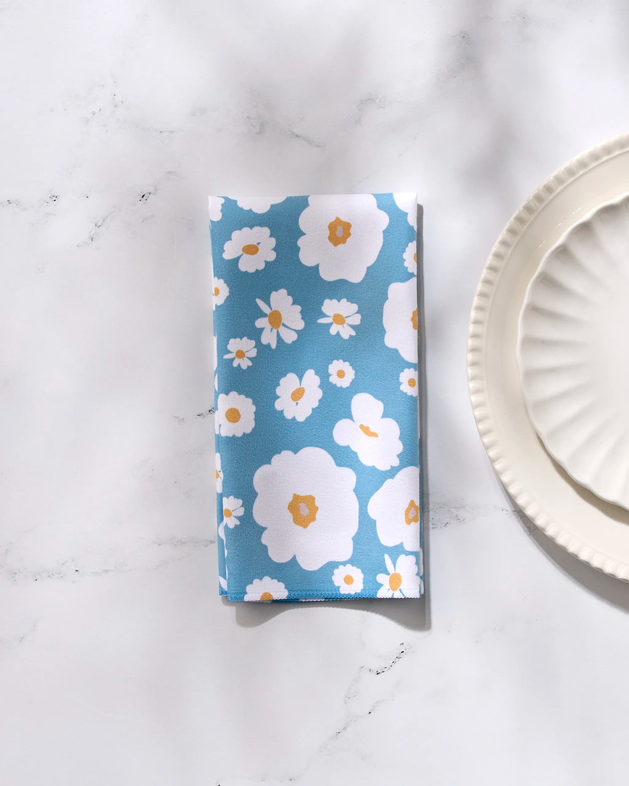 A GEOMETRY Happy Daysies blue dinner napkin, showcasing a white and yellow floral print, is neatly folded on a white marble surface beside two stacked scalloped white plates.