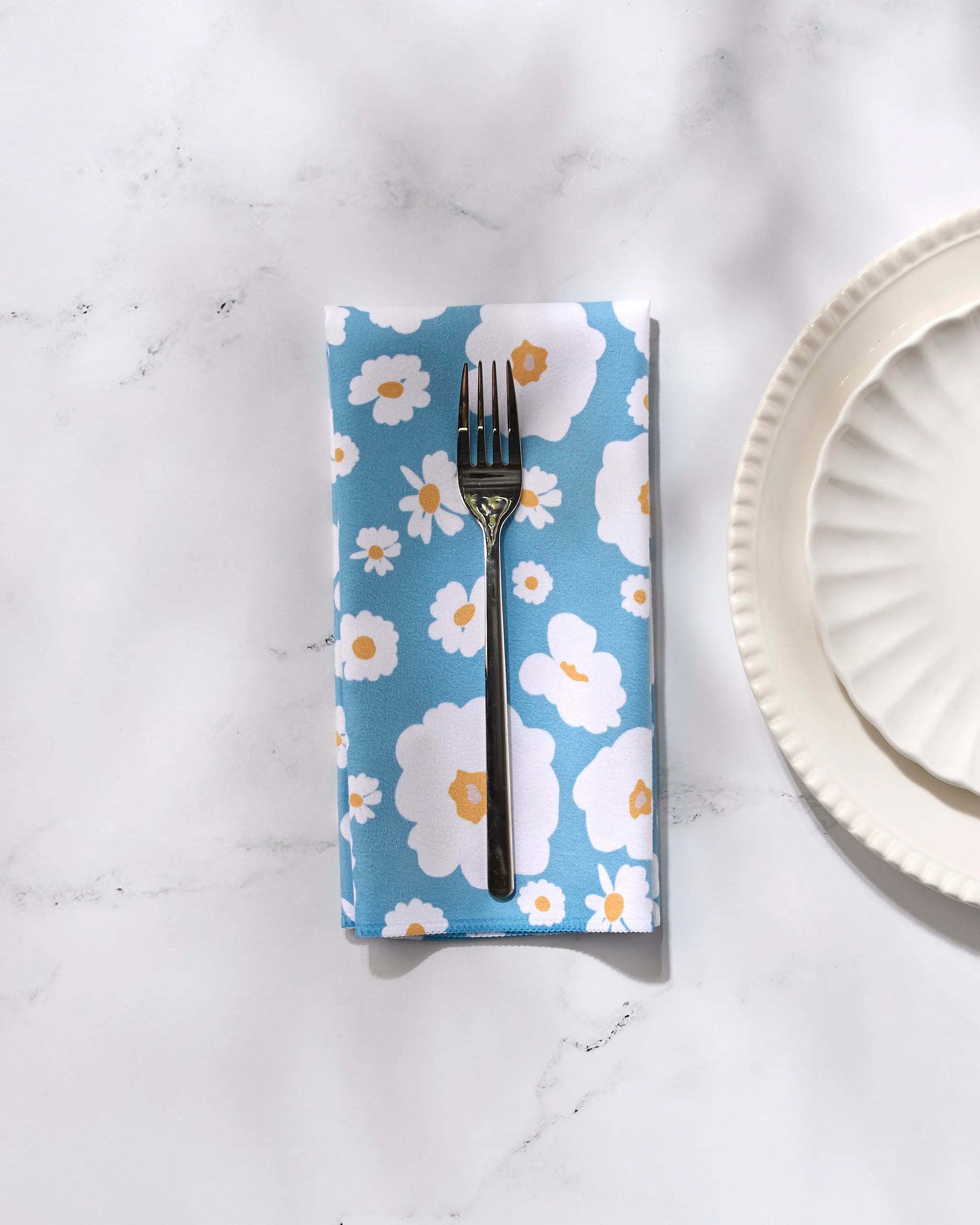 A fork rests on a GEOMETRY Happy Daysies Dinner Napkin Set with blue napkins featuring white and yellow flowers, crafted from reusable fabric, on a white marble surface next to two stacked, scalloped-edge plates.