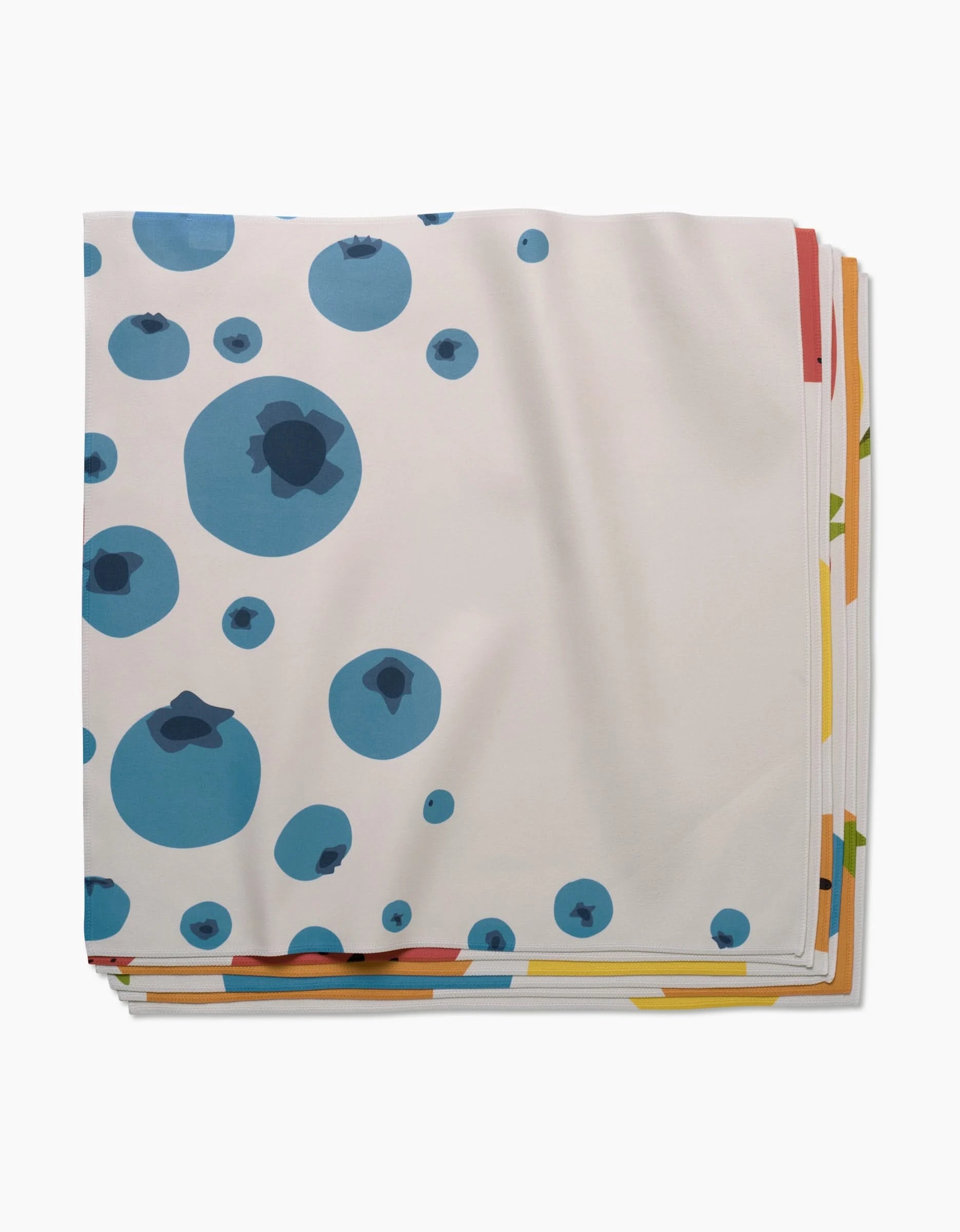 A folded white GEOMETRY Sunny Fruit dinner napkin with a blue blueberry-like circle pattern, made from Geosuede fabric, is stacked on colorful linens against a white background.