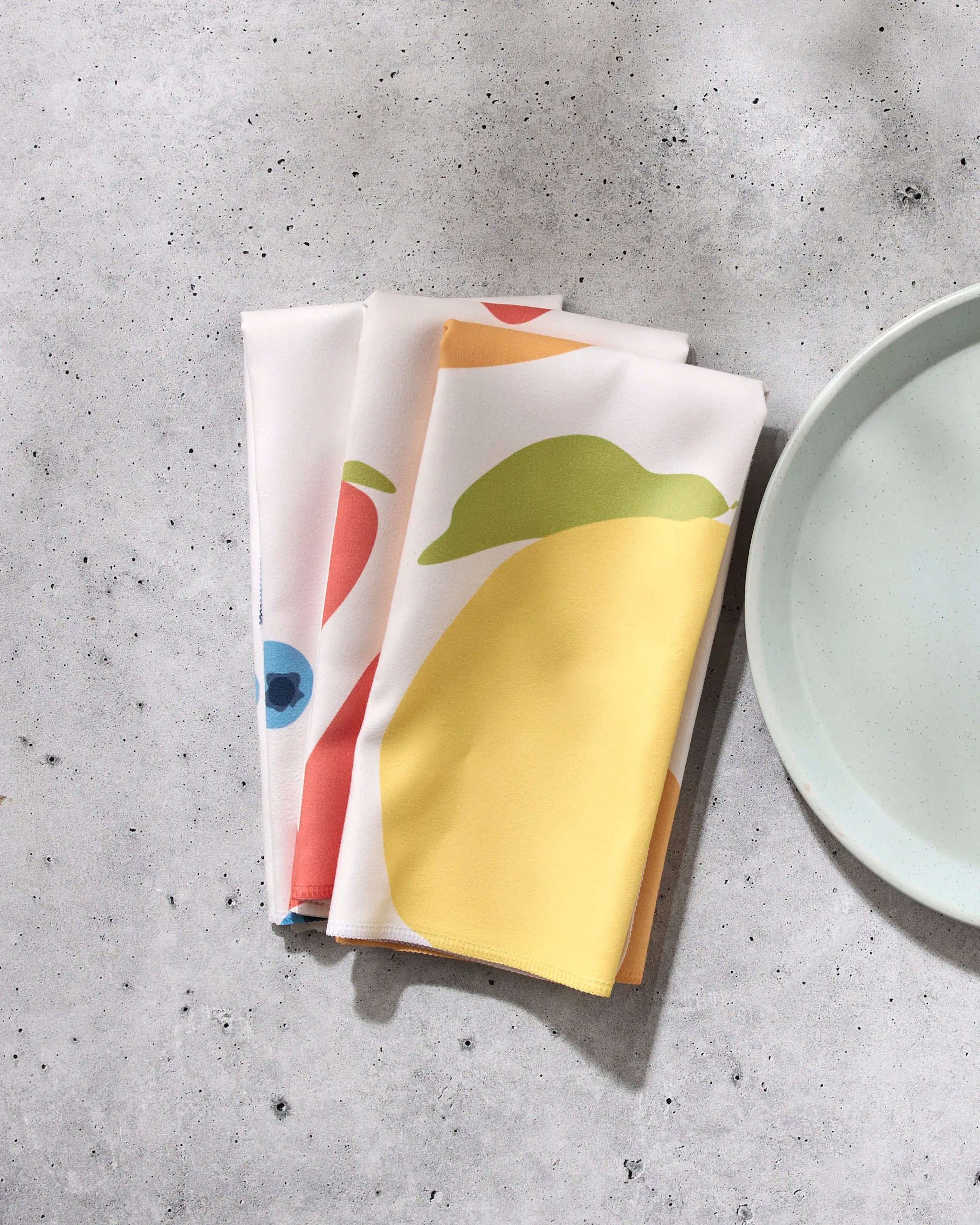 The GEOMETRY Sunny Fruit set features four folded white reusable dinner napkins with vibrant yellow, green, red, and blue fruit prints, displayed on a light gray speckled surface beside a pale blue plate.