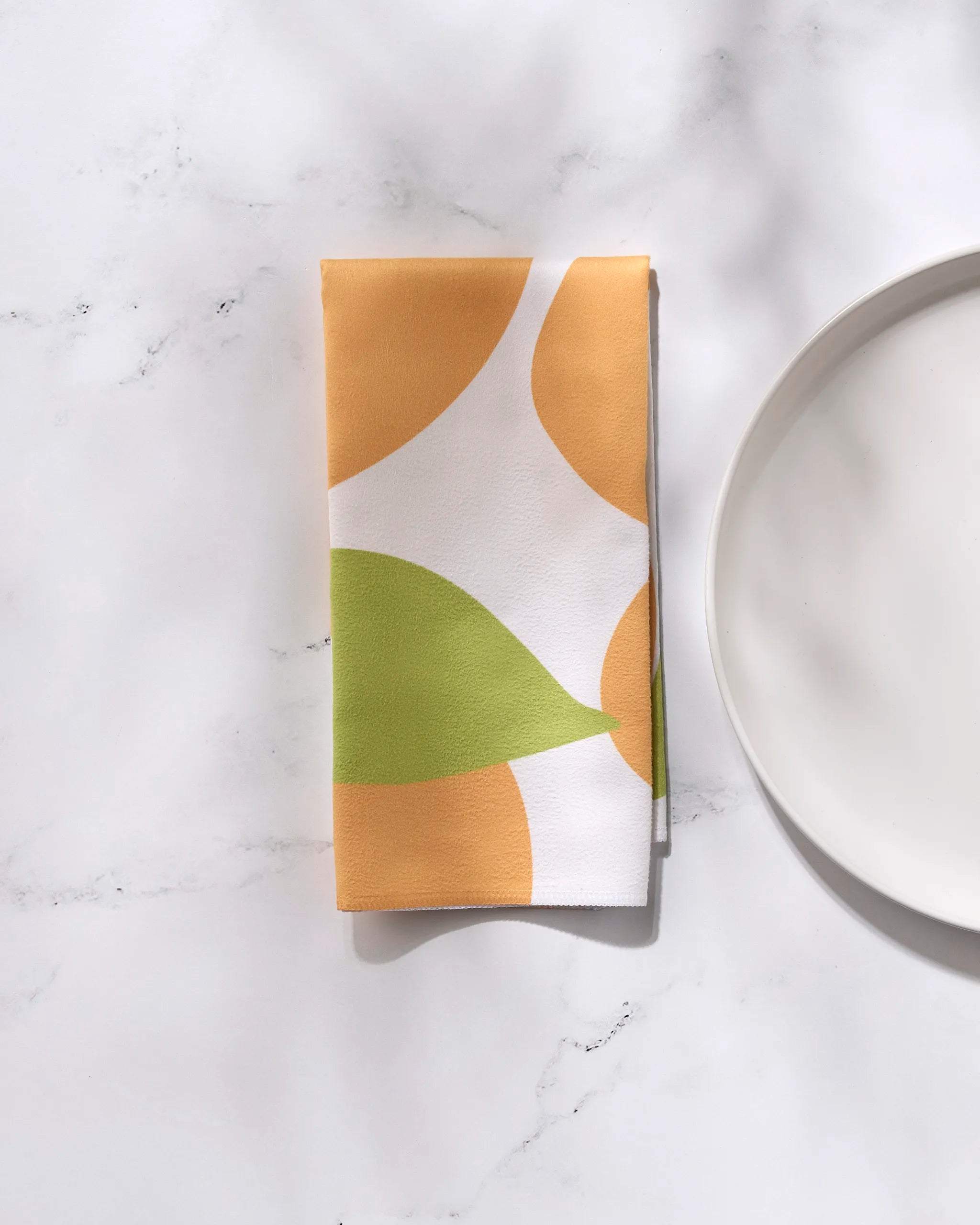 A folded GEOMETRY "Sunny Fruit" reusable dinner napkin with an abstract orange and green pattern rests beside a plain white plate on a white marble surface.