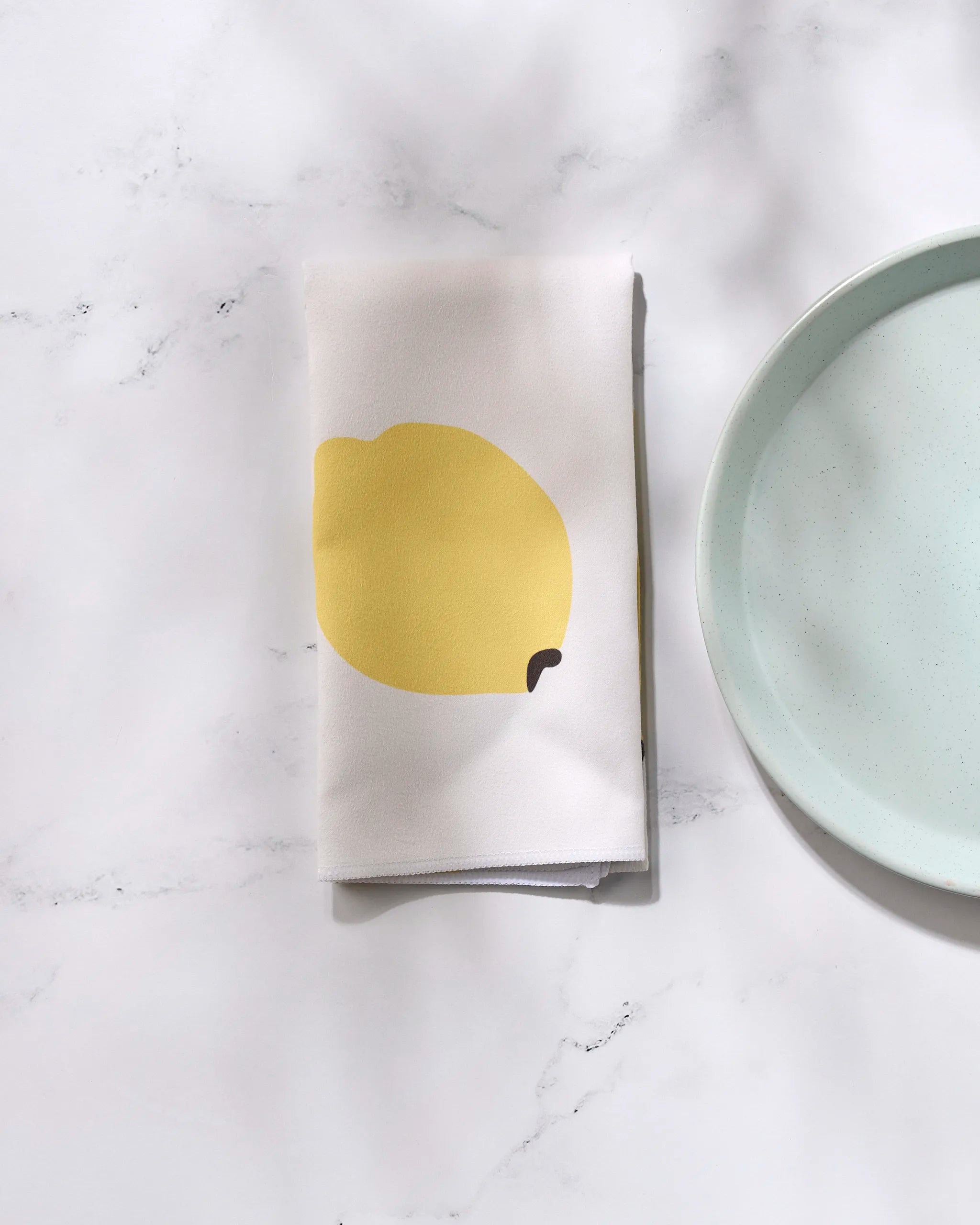 The GEOMETRY "Sunny Fruit" folded white reusable dinner napkin with a yellow lemon illustration sits on a white marble surface beside a light blue plate.