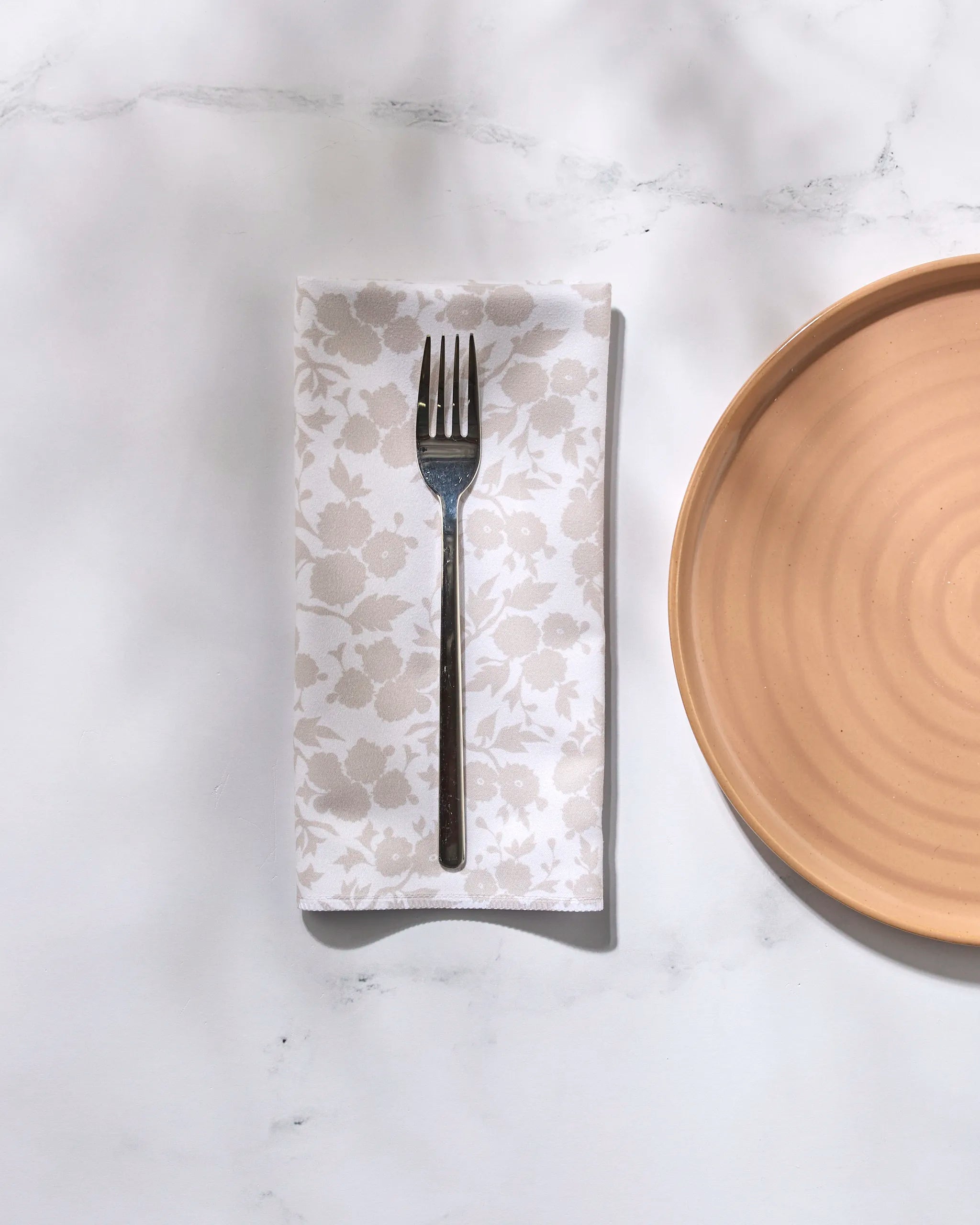 A metal fork rests on a folded GEOMETRY Betty's Buds floral-patterned recycled dinner napkin beside a round tan ceramic plate on a white marble surface with soft shadows.