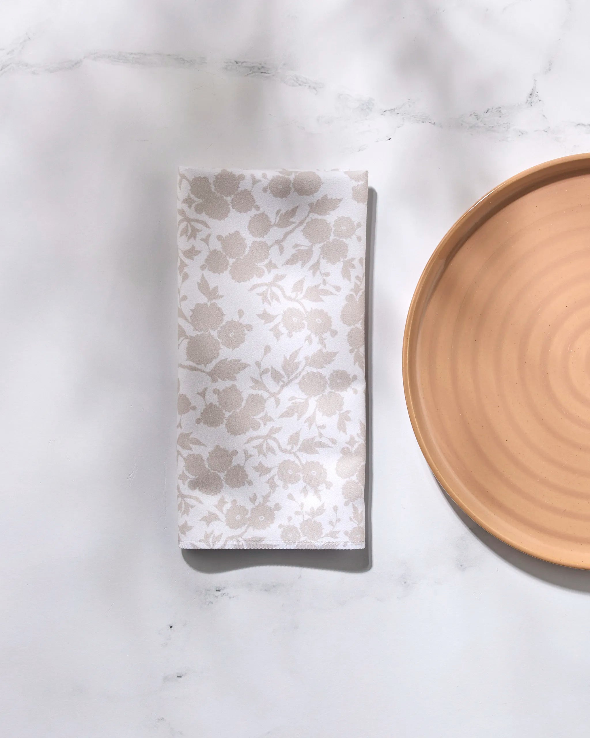 The GEOMETRY "Betty's Buds" beige ceramic plate with concentric circles sits on a white marble surface beside a folded recycled dinner napkin featuring a subtle floral pattern.