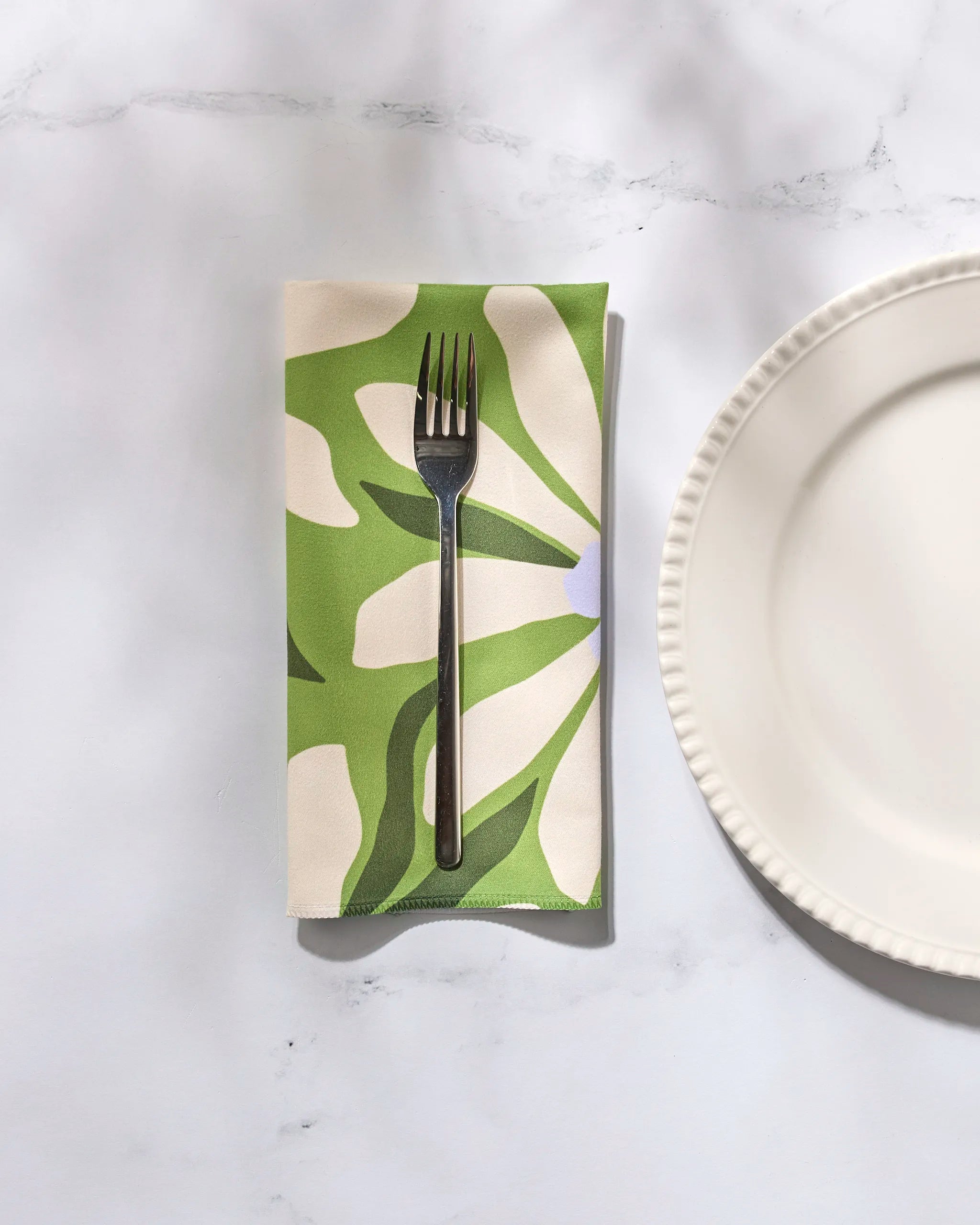A metal fork rests on a Bliss and Bloom Dinner Napkin from GEOMETRY, featuring a green and white pattern, beside a white scalloped plate—all arranged on a light-colored surface.