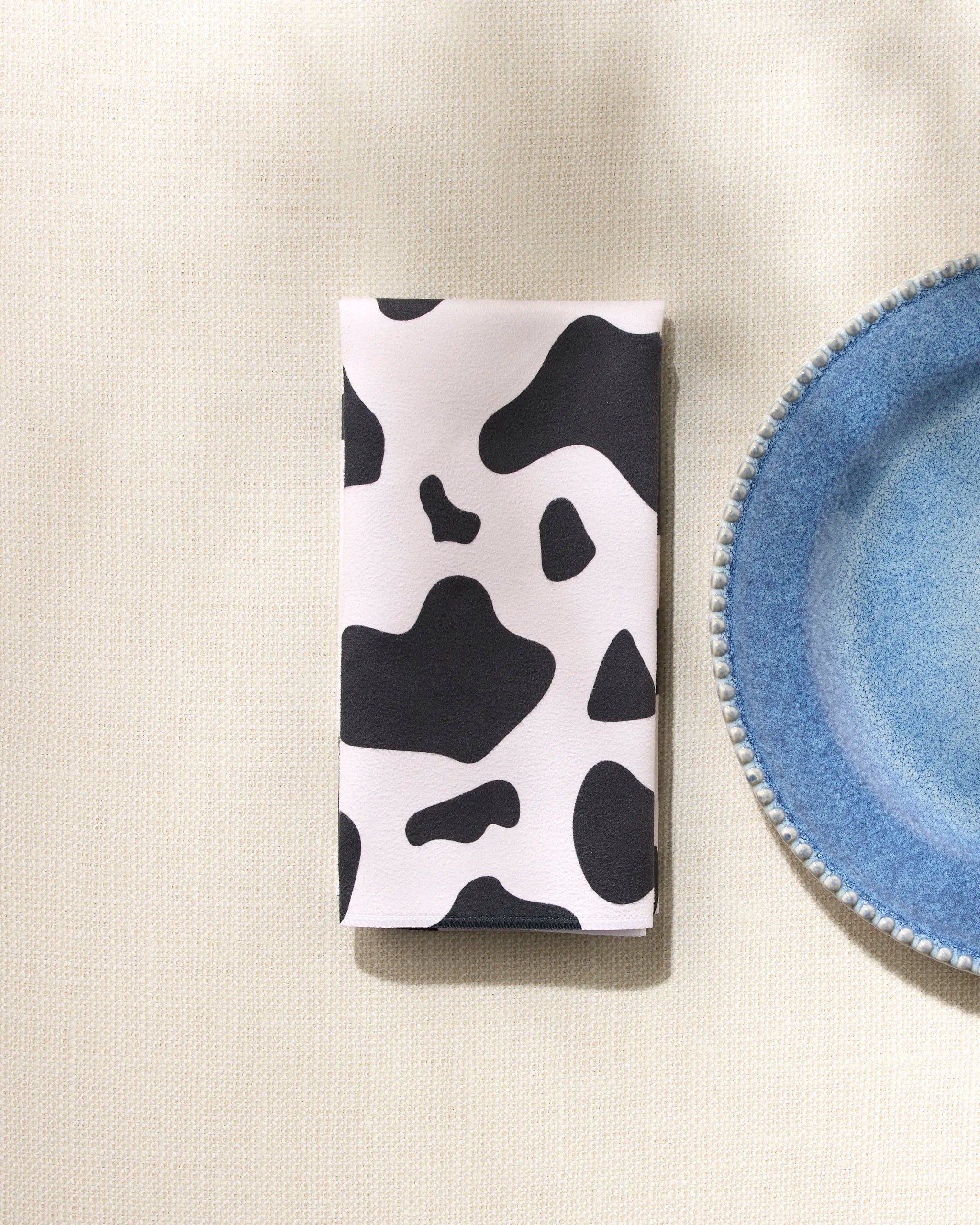 A folded GEOMETRY Cow Print Black eco-friendly napkin rests beside a blue textured-rim ceramic plate on a light, textured surface.