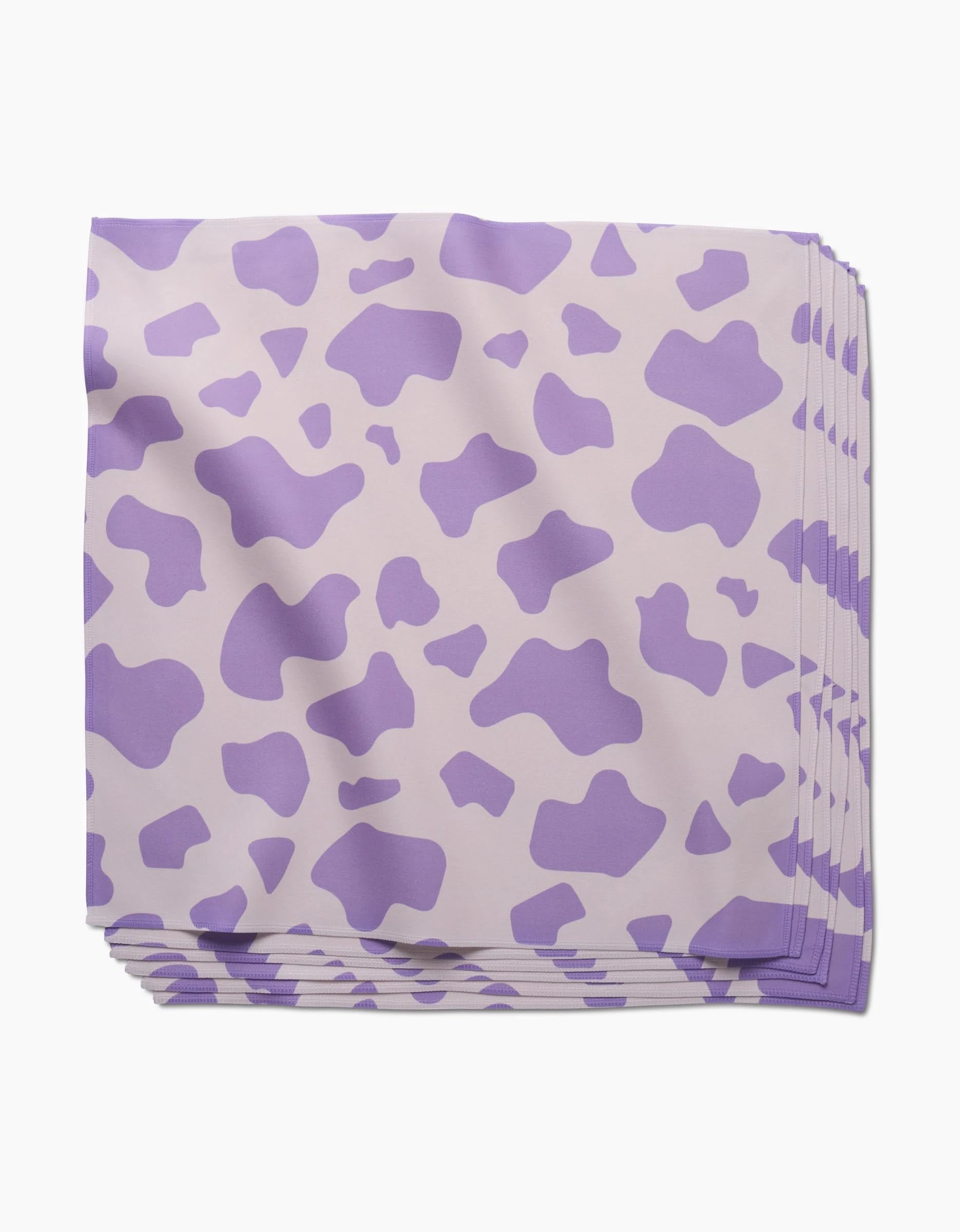 A neatly stacked set of GEOMETRY Cow Print Lilac absorbent cloth napkins featuring a light purple cow-print pattern on a pale background, displayed on a white surface.