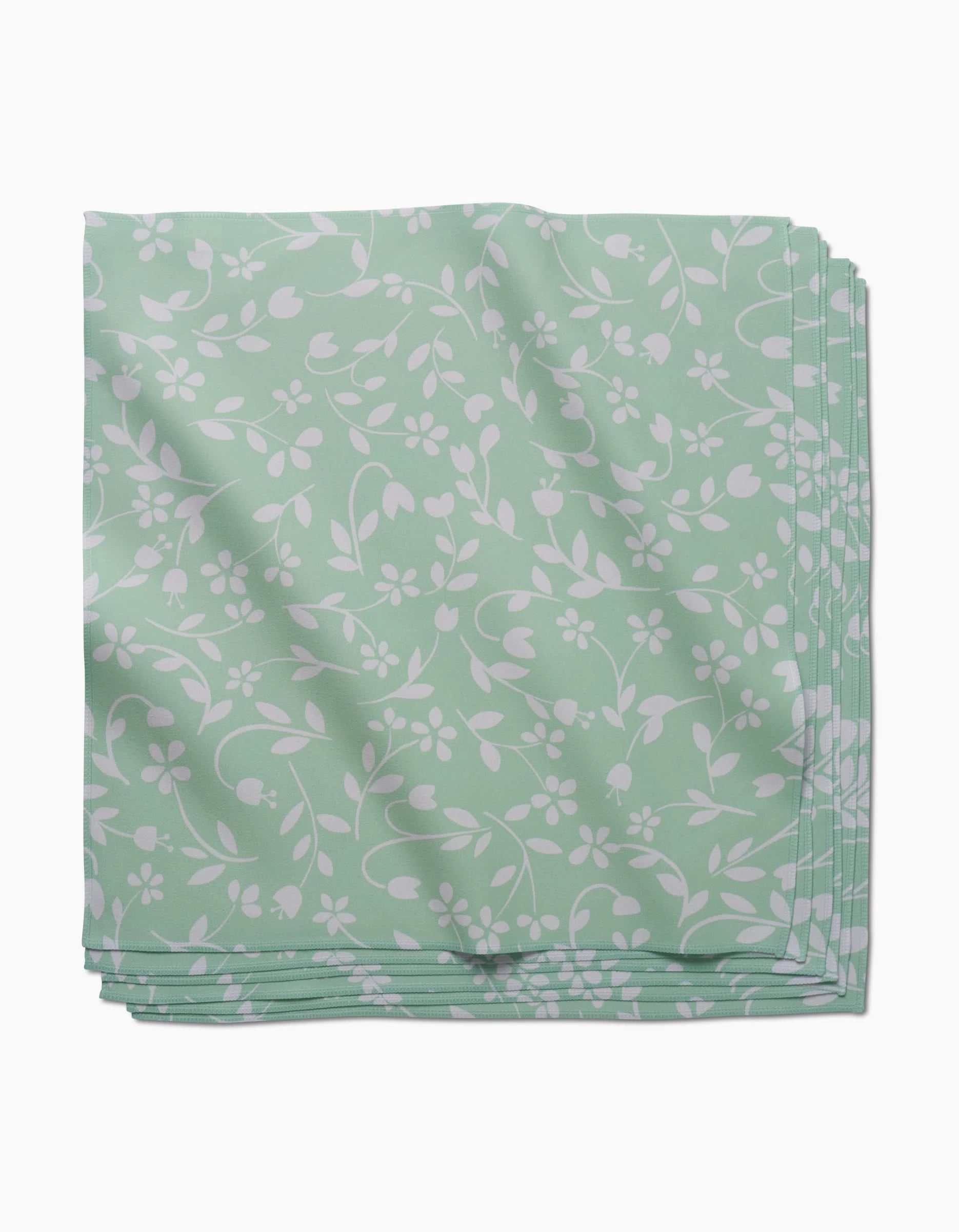 A neatly folded stack of GEOMETRY Dandy light green absorbent cloth napkins with a white floral and vine pattern, displayed on a white background.