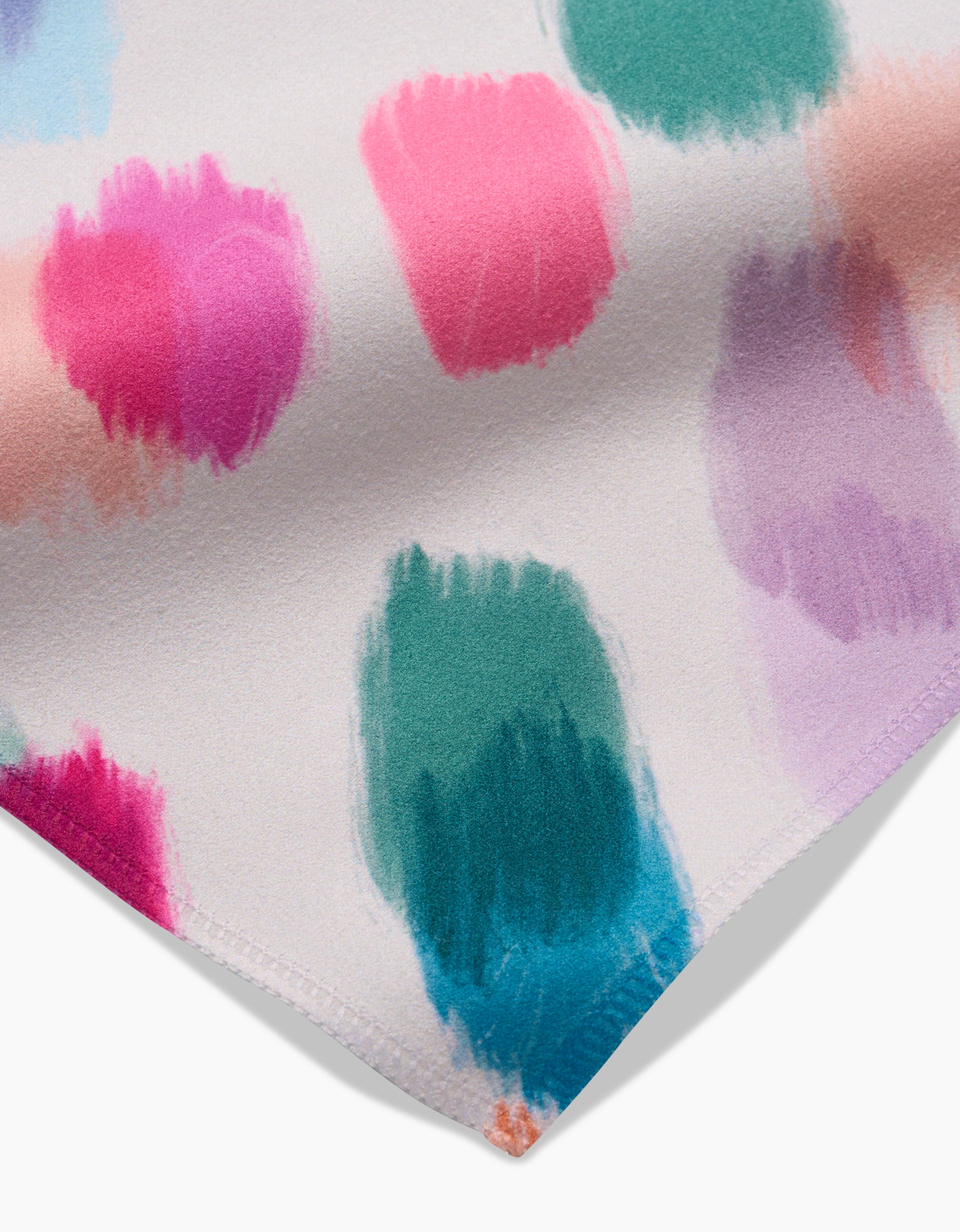 A close-up of GEOMETRYs Downpour Geosuede Dinner Napkins, made from recycled materials, showcases colorful brushstroke spots in pink, green, blue, and purple on white with neatly hemmed corners.